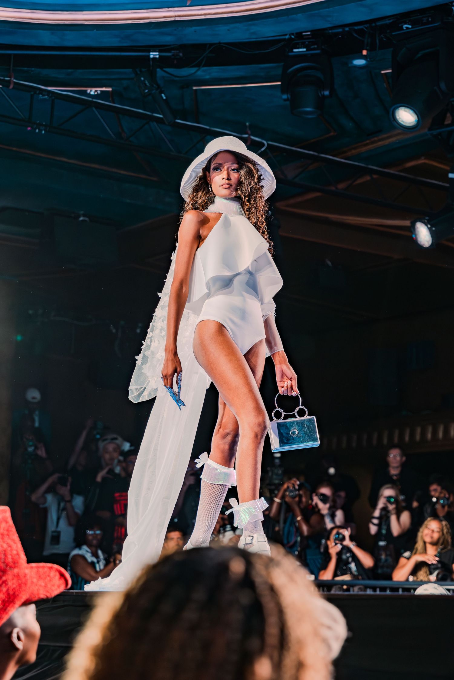 Model walks runway in white asymmetric dress, bucket hat, mini bag, and bow socks at a fashion show.