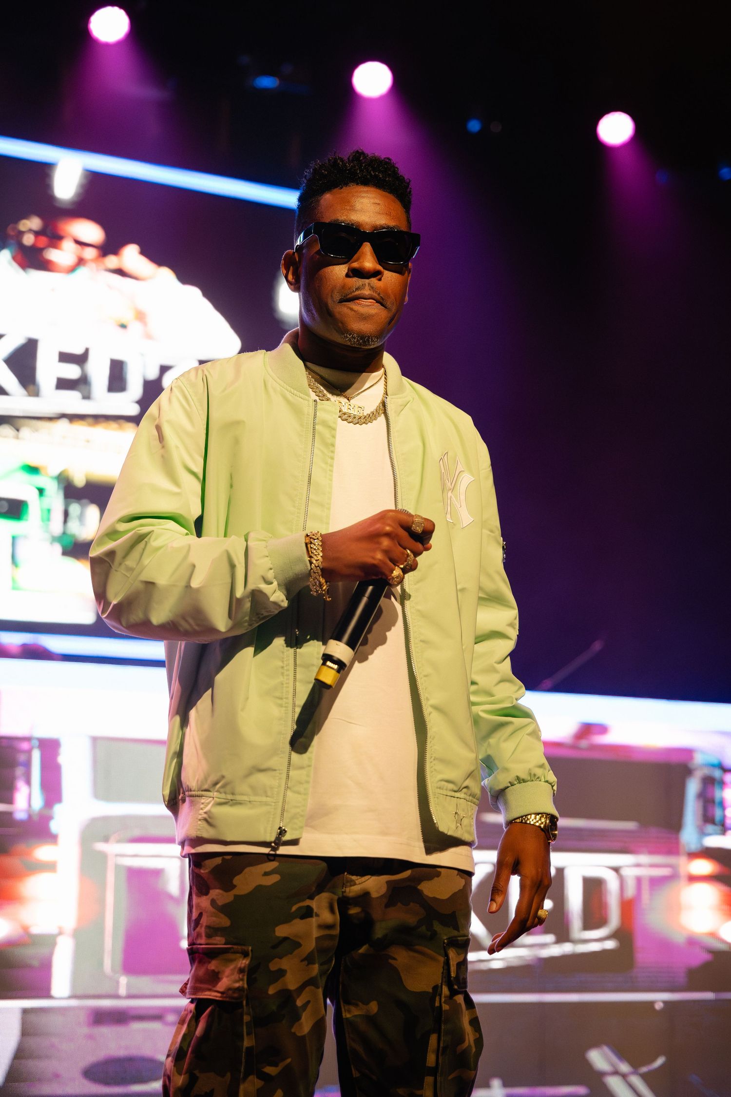 Male performer in green bomber jacket and camo pants holding microphone on stage.