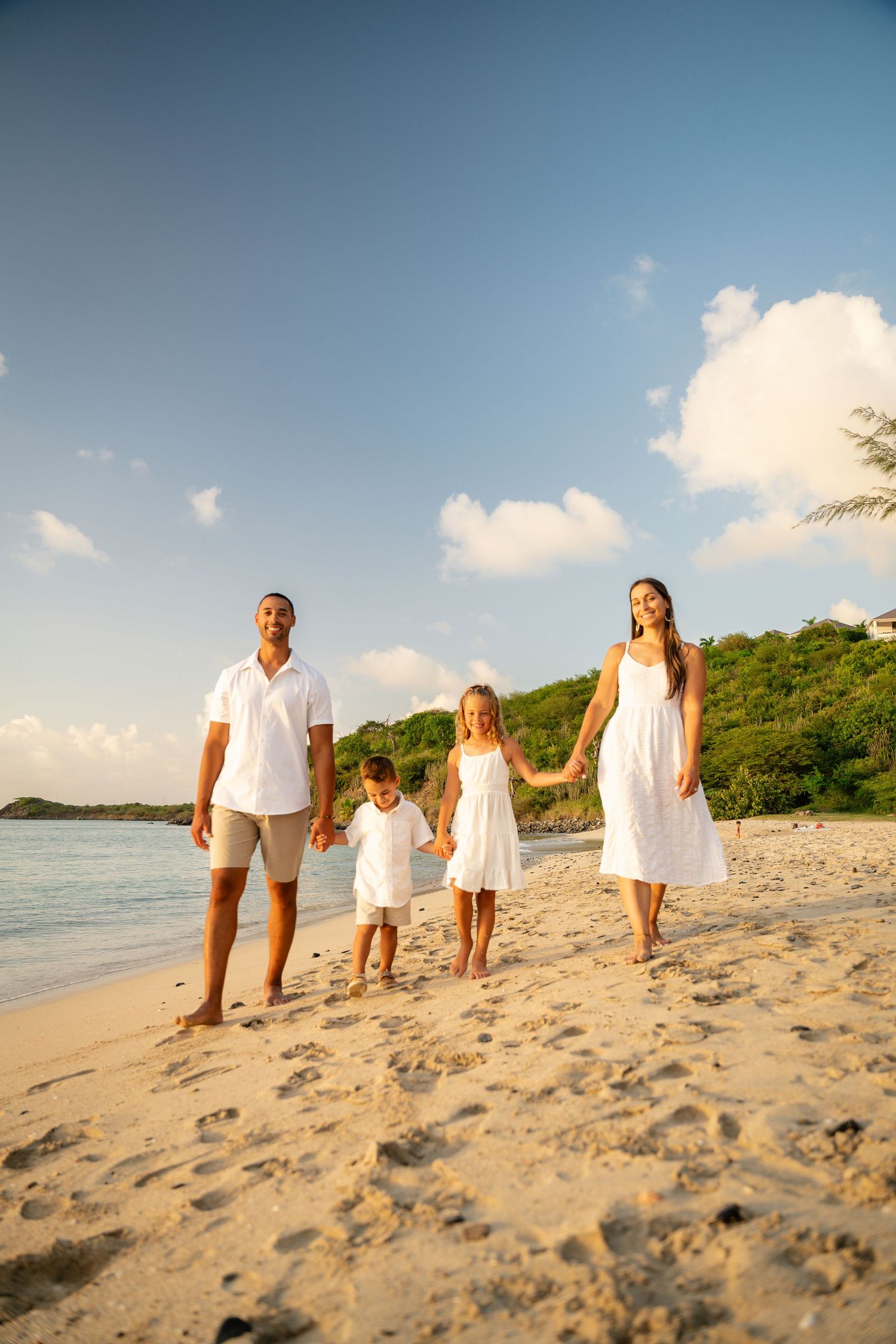 Happy family of four in white outfits holding hands while walking along a sunny tropical beach.