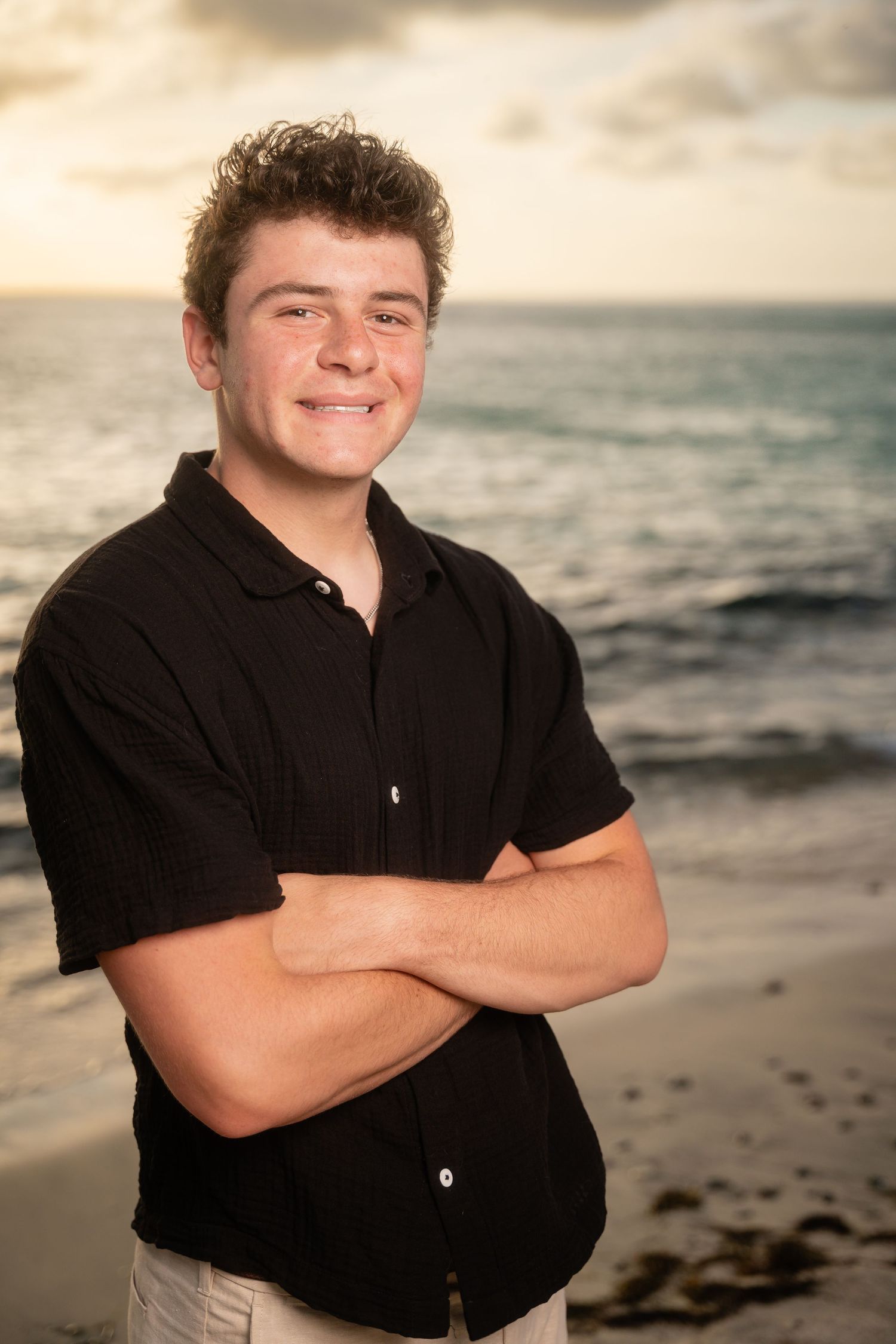 Smiling teenage boy in black shirt with arms crossed posing on beach at sunset.