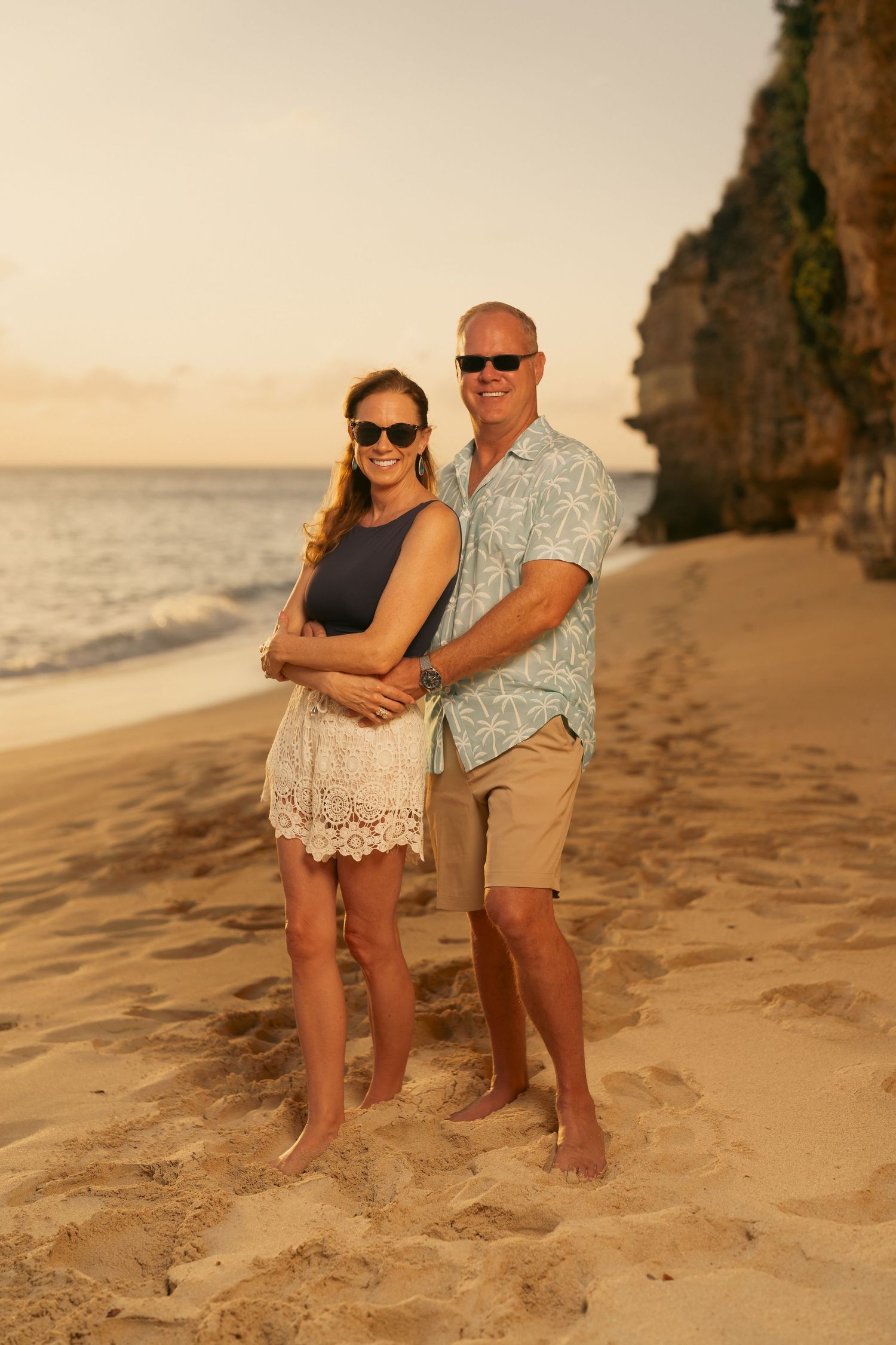 Smiling couple in sunglasses embracing on tropical beach at sunset near rocky cliffs.