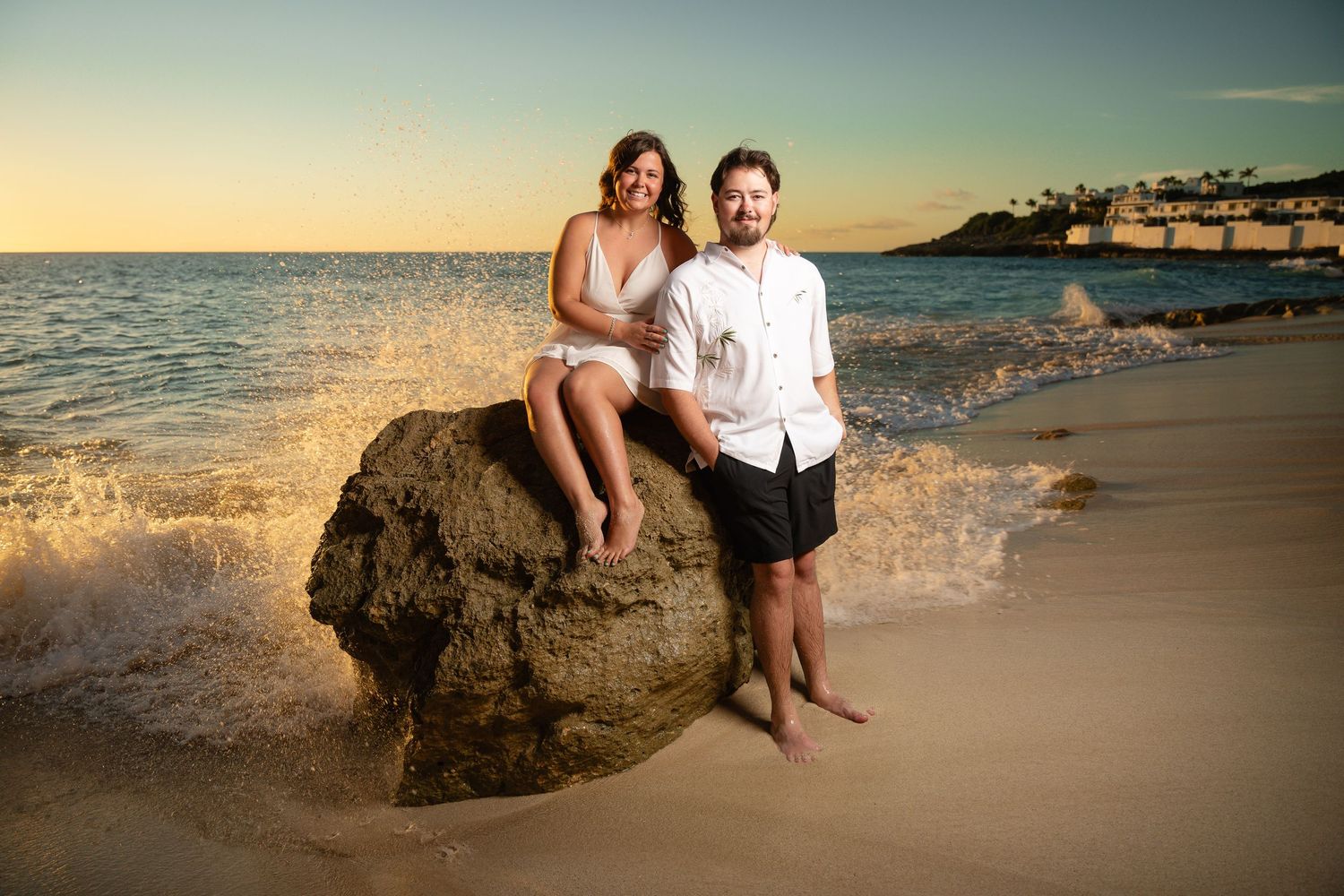 Couple posing on a large rock at the beach during sunset, woman sitting on rock beside standing man with waves splashing.