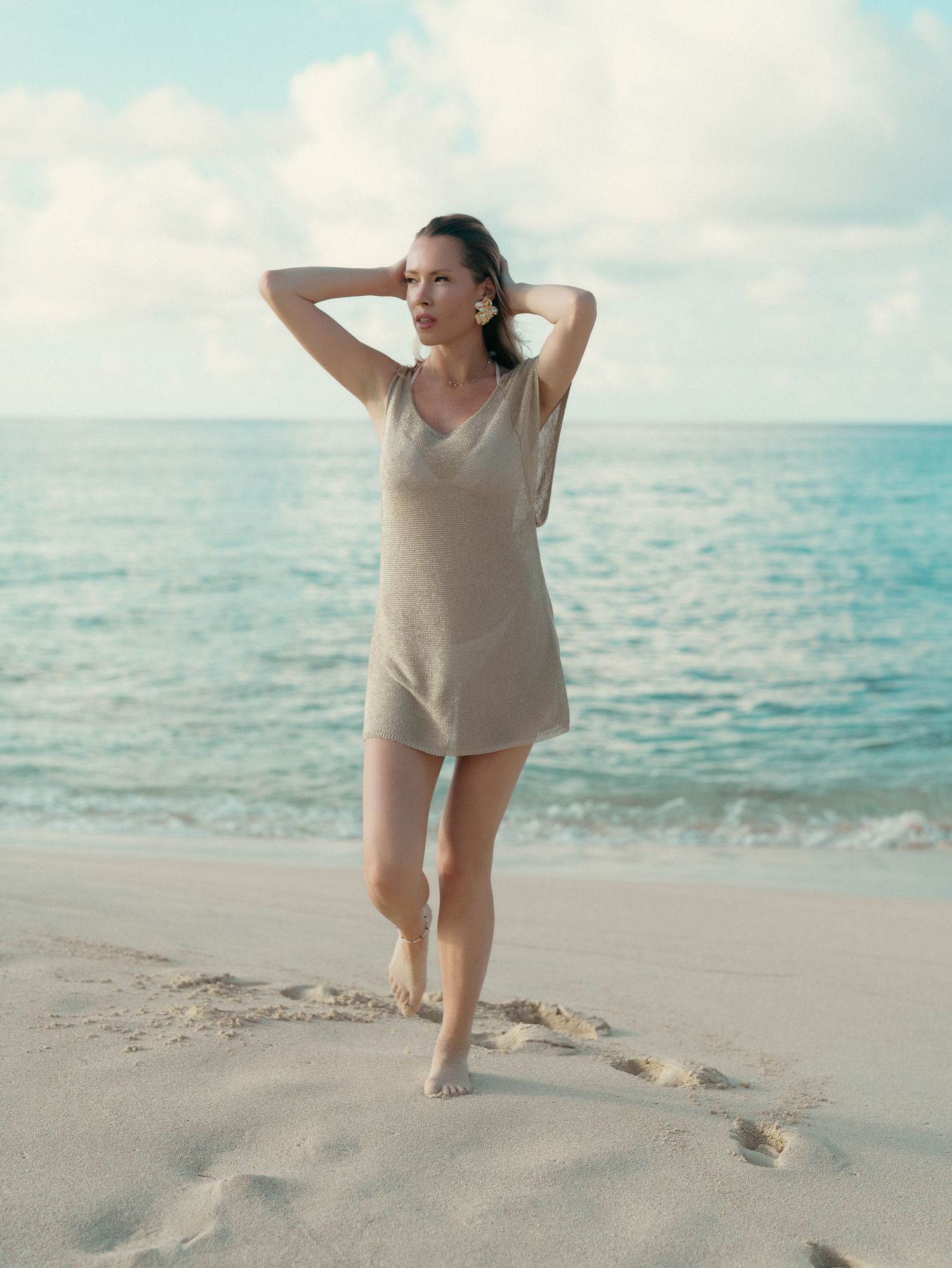 Slender woman in beige knit mini dress posing barefoot on tropical sandy beach.
