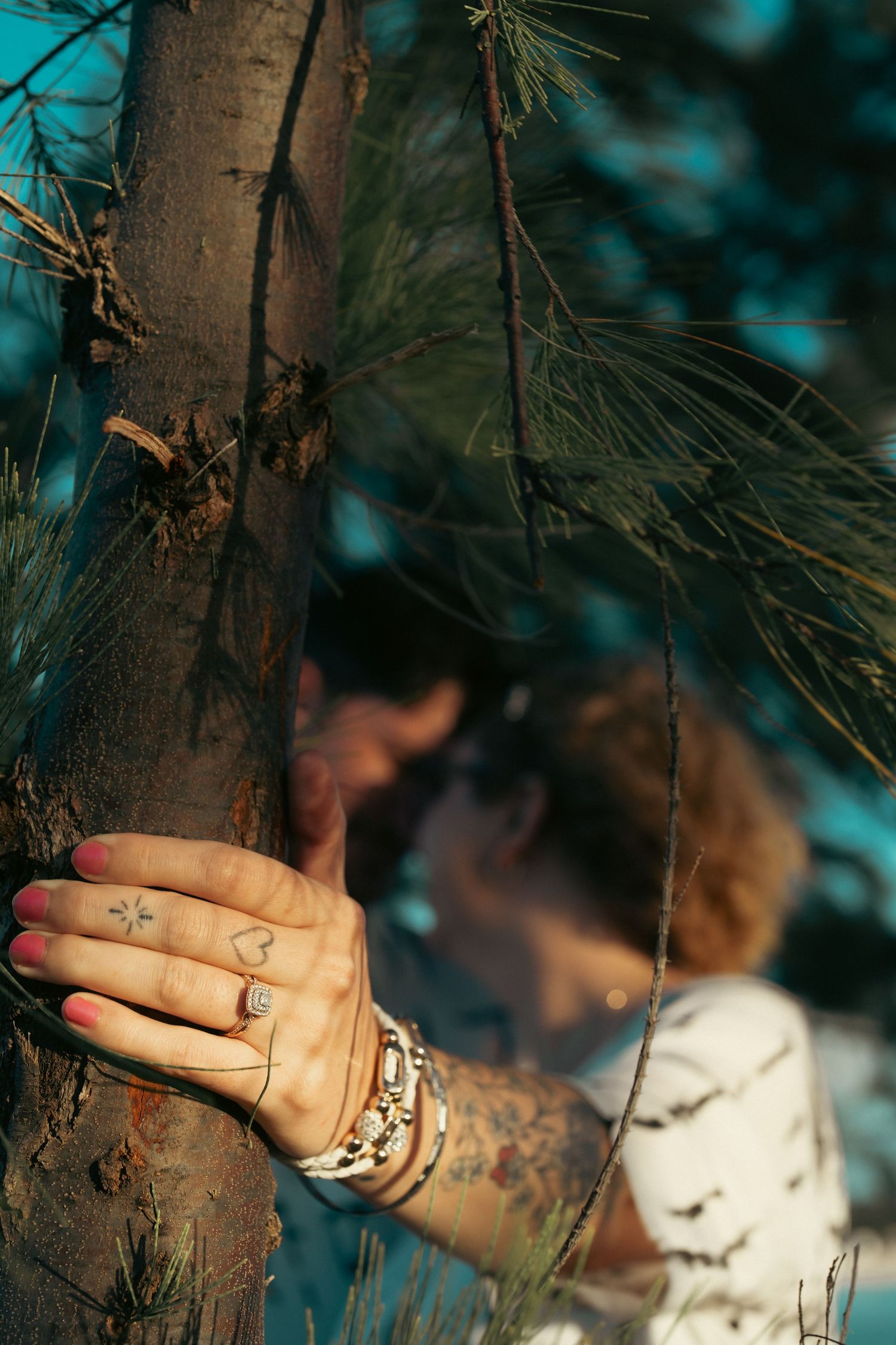 Woman's tattooed hand with ring resting on pine tree trunk while couple kisses behind.