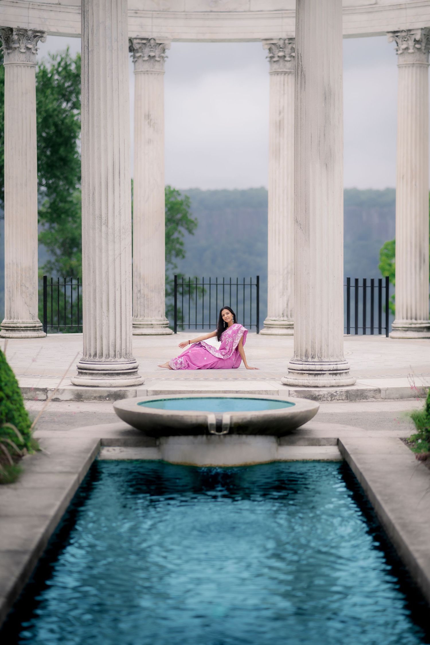 Woman in pink saree sitting near fountain surrounded by classical white columns outdoors.