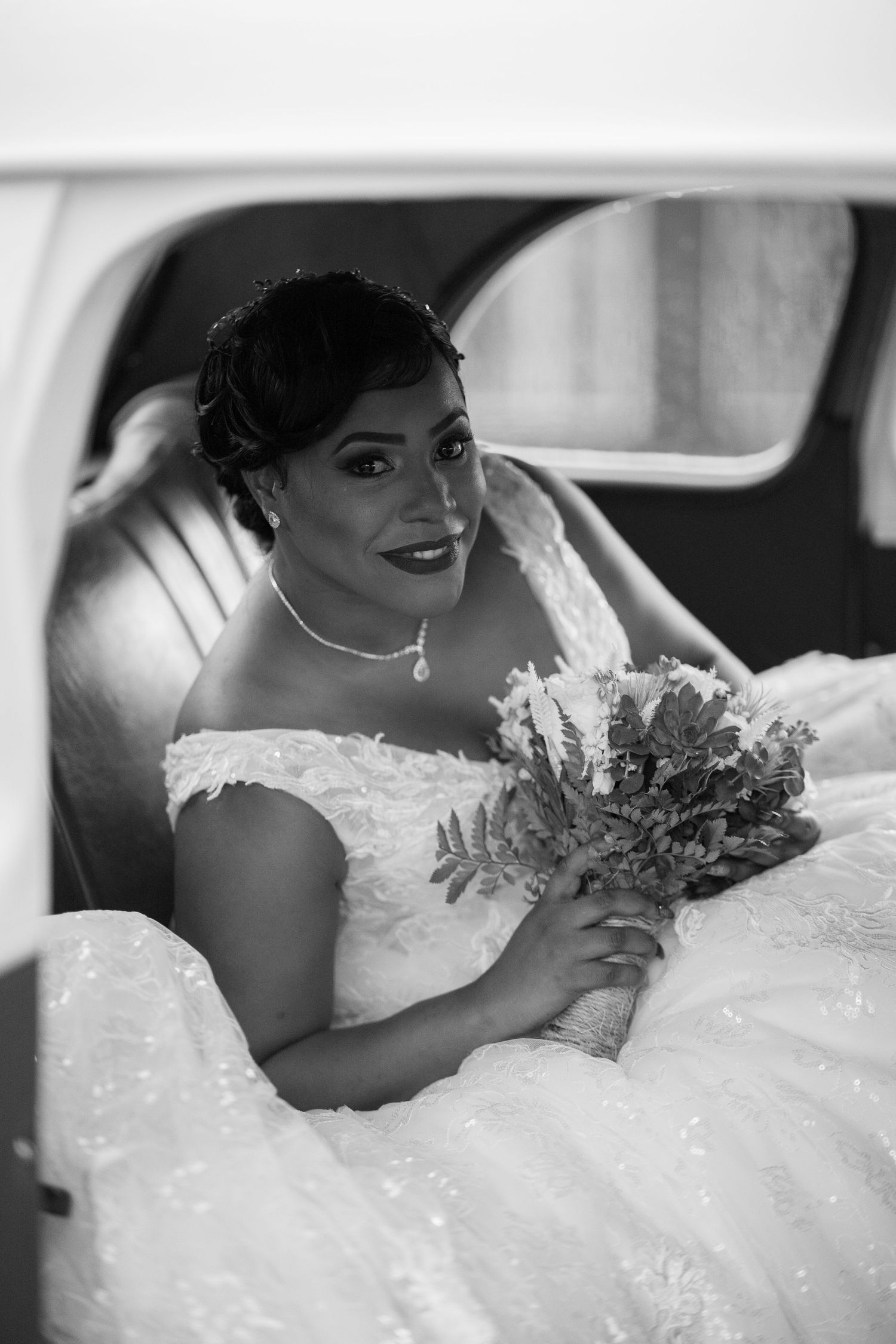 Elegant bride in lace gown holds bouquet while seated in a car on her wedding day.
