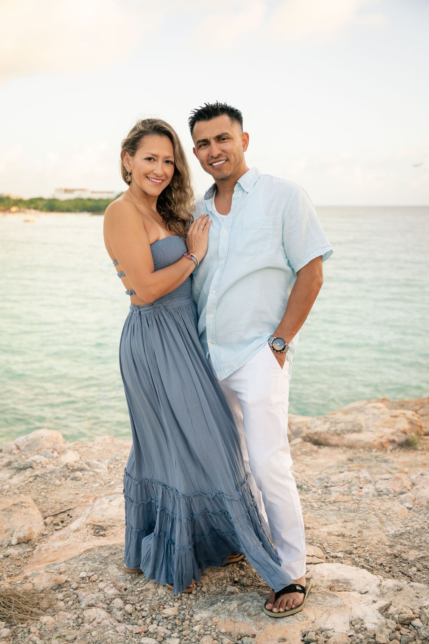 A smiling couple poses together on rocky oceanfront, woman in blue dress and man in light blue shirt.