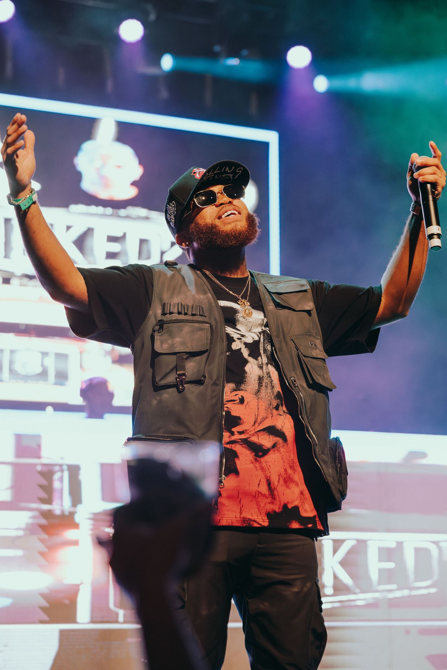 A bearded male artiste raises his arms on stage before a lit backdrop at a live concert.