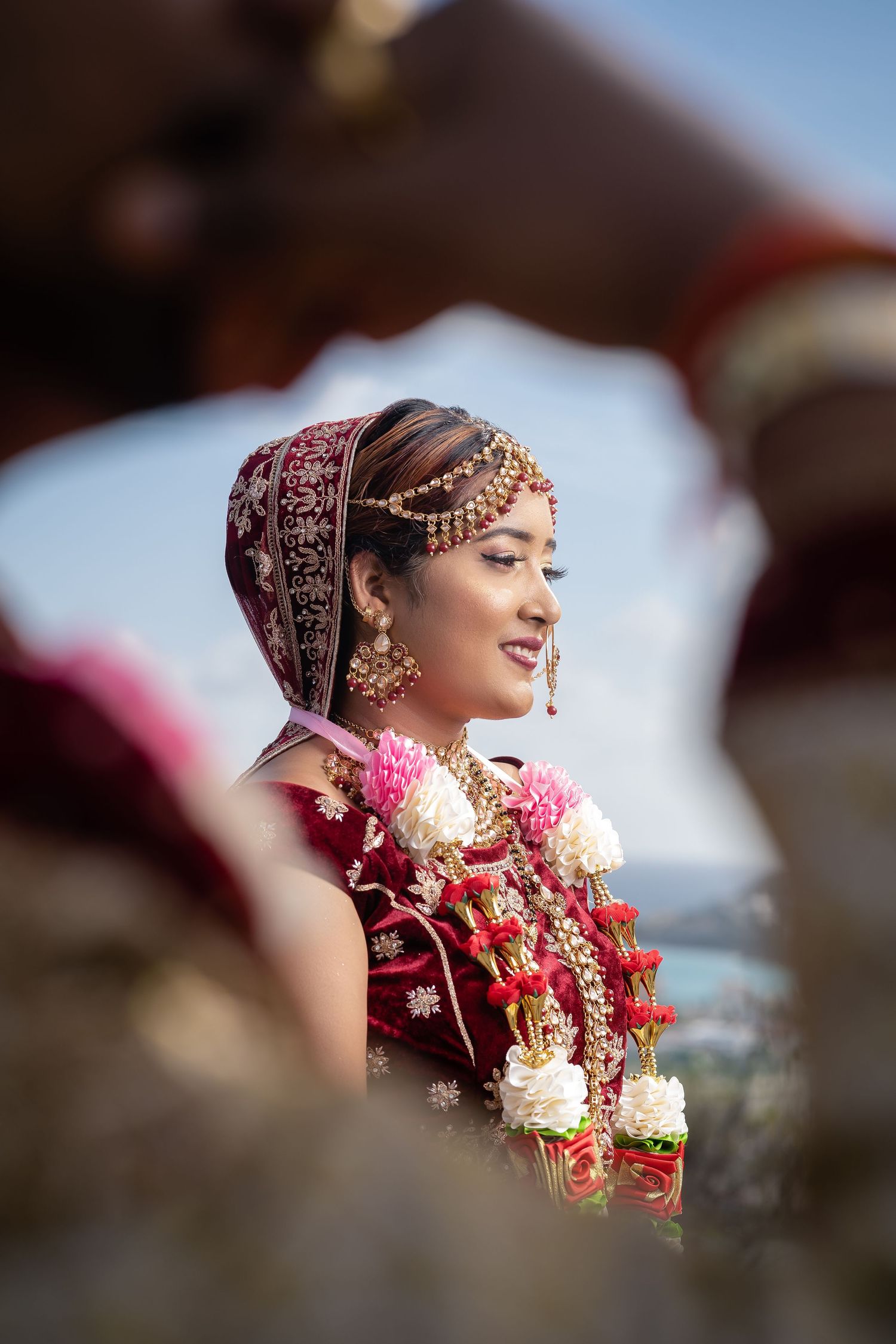 Indian bride in red bridal attire with gold jewelry and floral garlands at wedding.