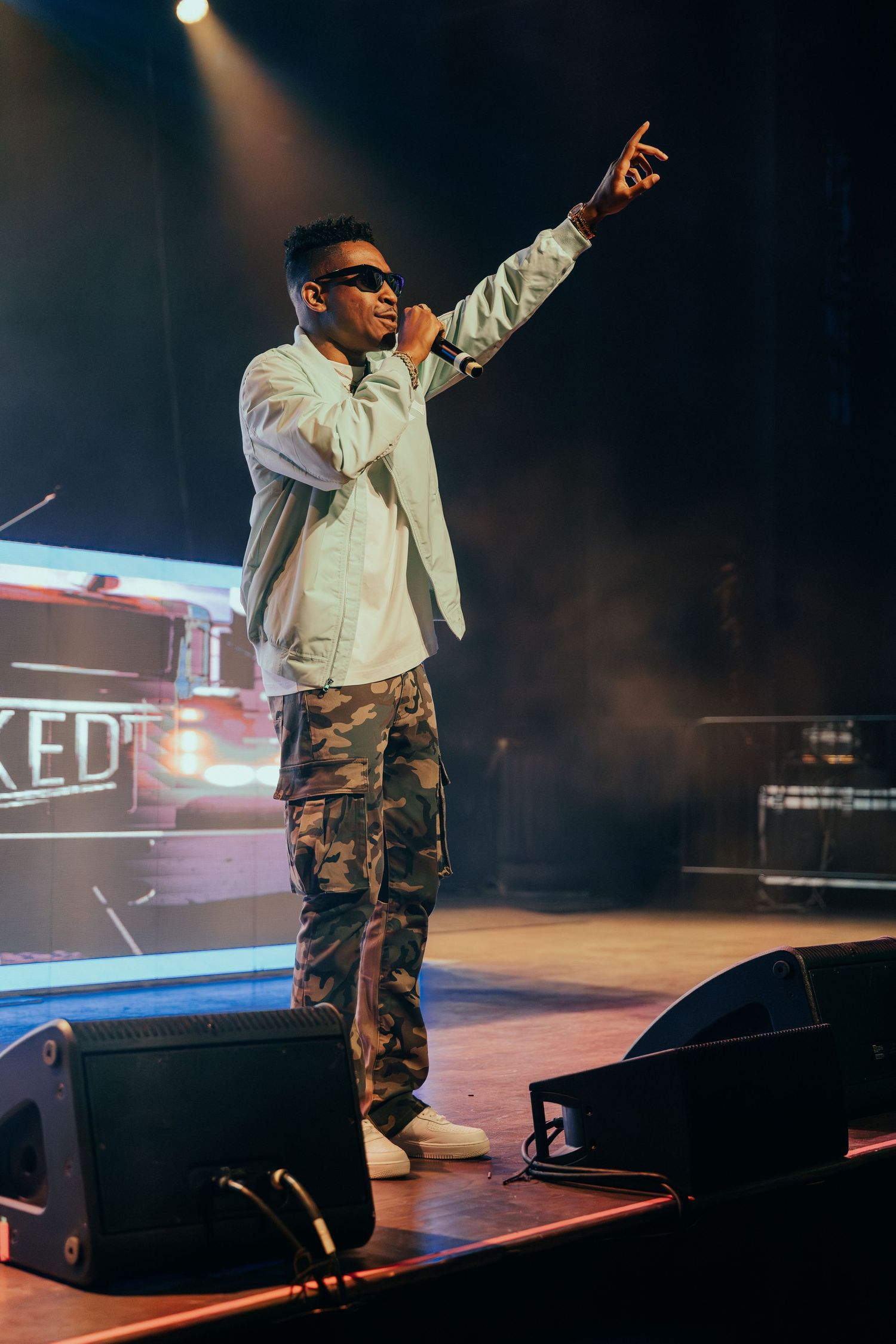 Male performer in camo pants pointing to crowd while performing on a concert stage.