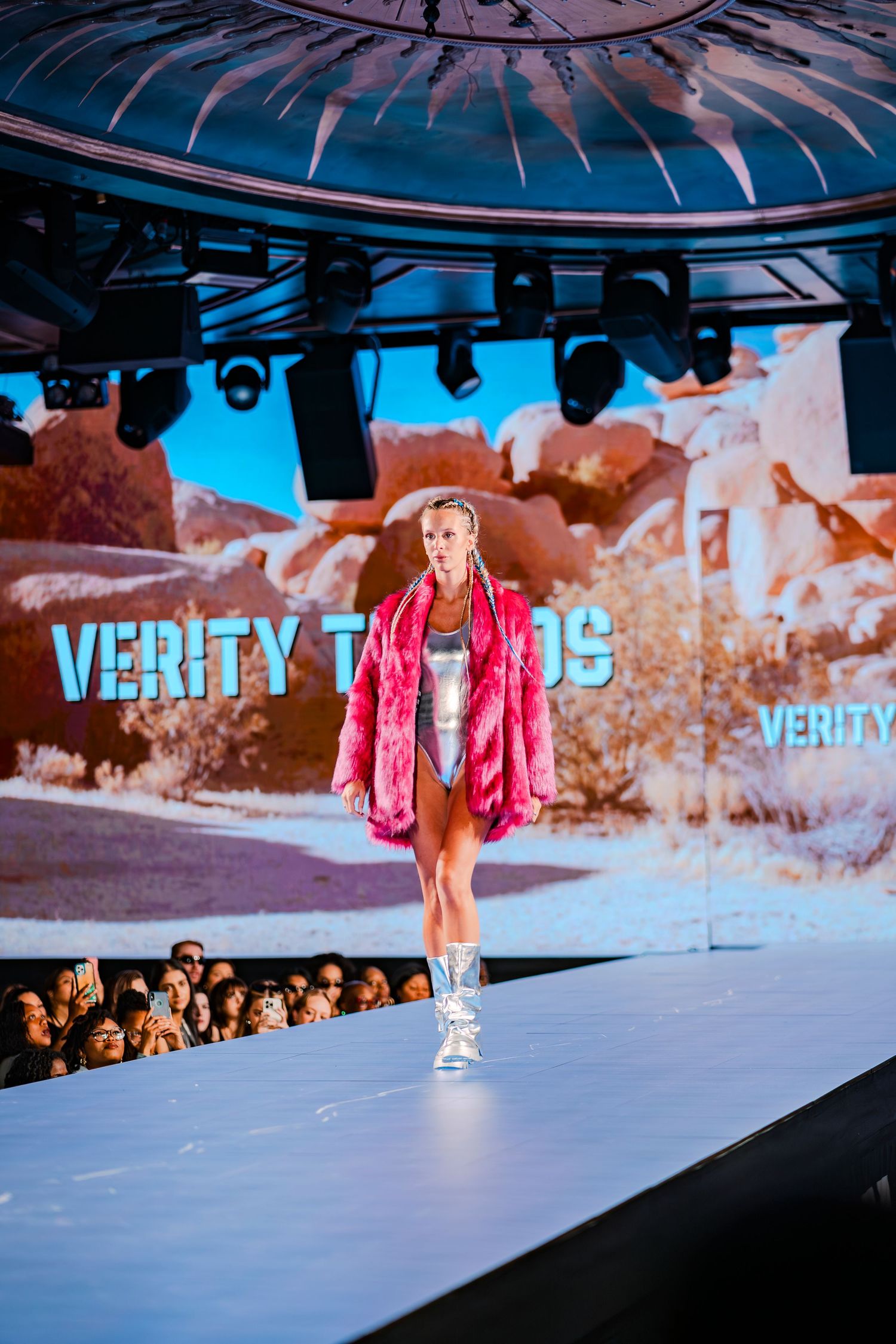 Model walks runway in pink fur coat at Verity Trends fashion show with desert backdrop.