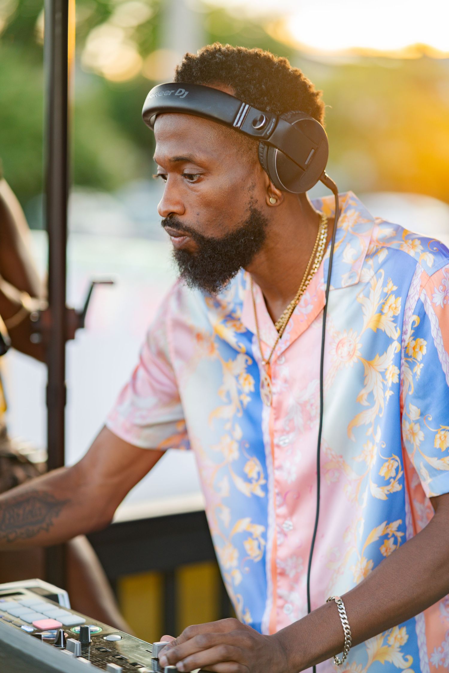 Bearded DJ wearing headphones and a floral shirt mixing music at an outdoor sunset event.