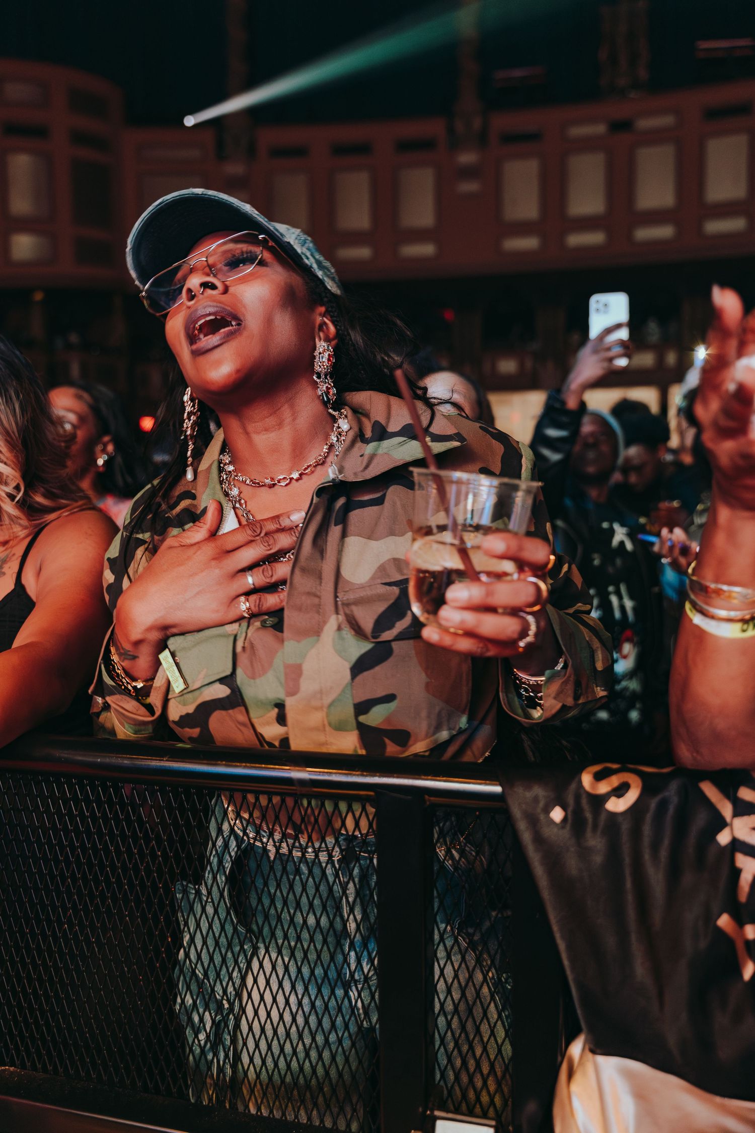 Woman in camo jacket and cap holding drink and singing along at a live concert.