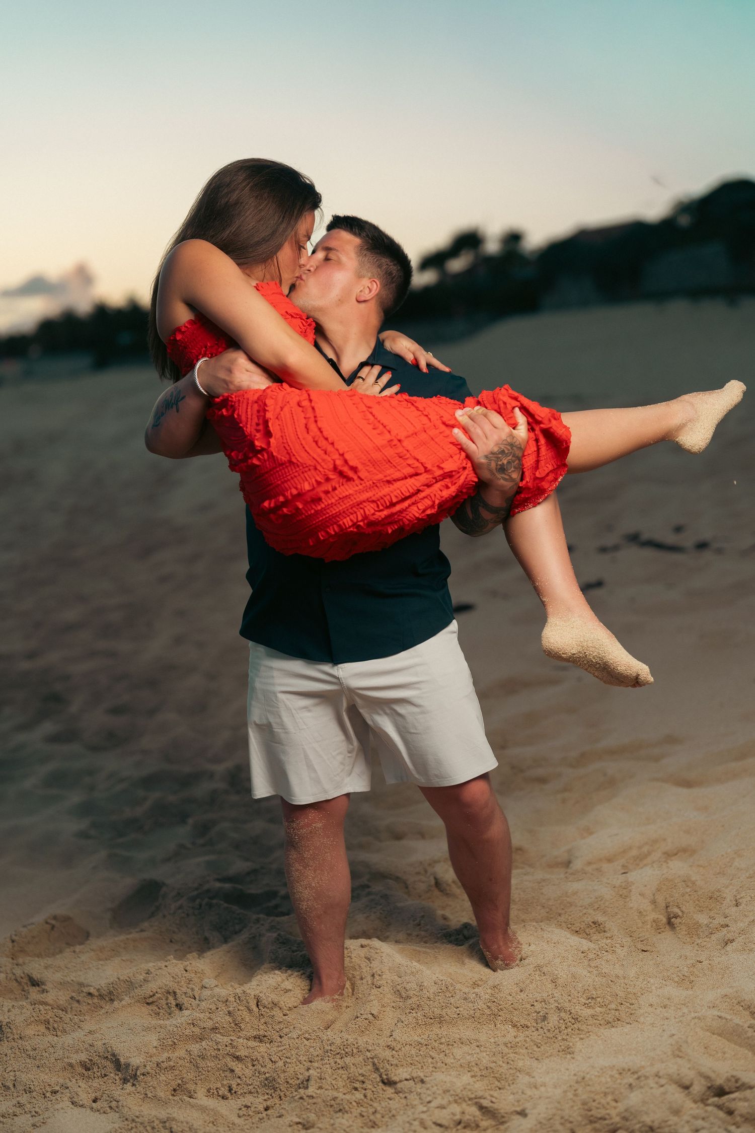 Man lifting and kissing a woman in a red dress on a sandy beach at dusk.