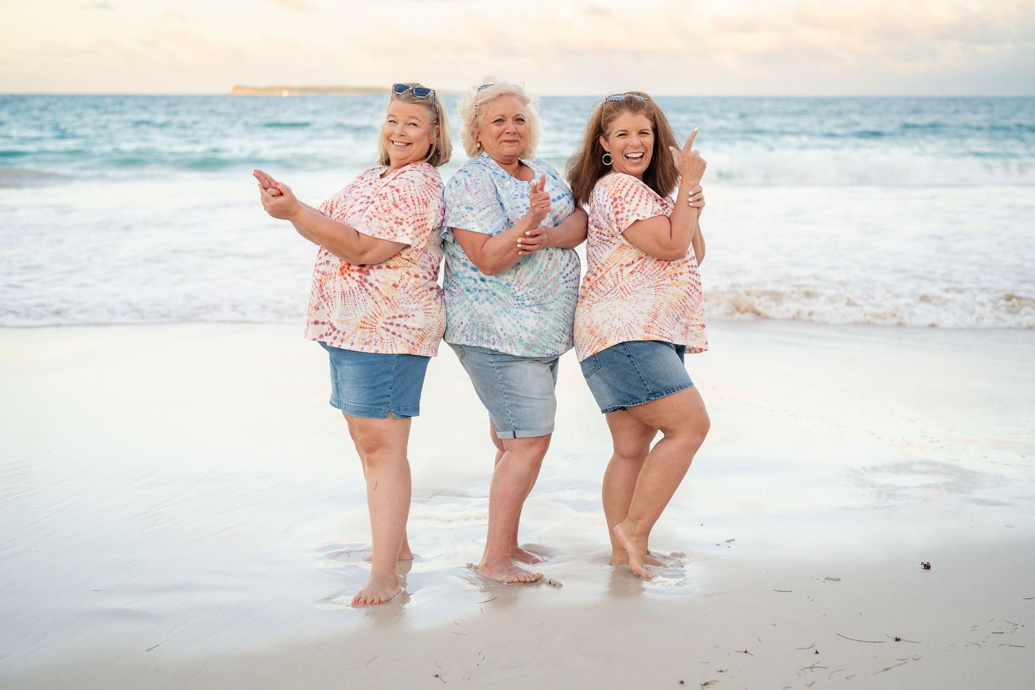 Three women in matching floral tops and denim skirts posing playfully together at the ocean's edge.