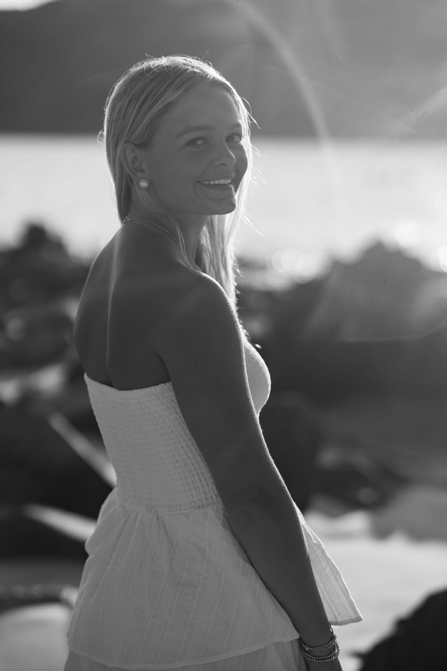 Black and white photo of smiling blonde woman in strapless white dress on rocky sunlit beach.