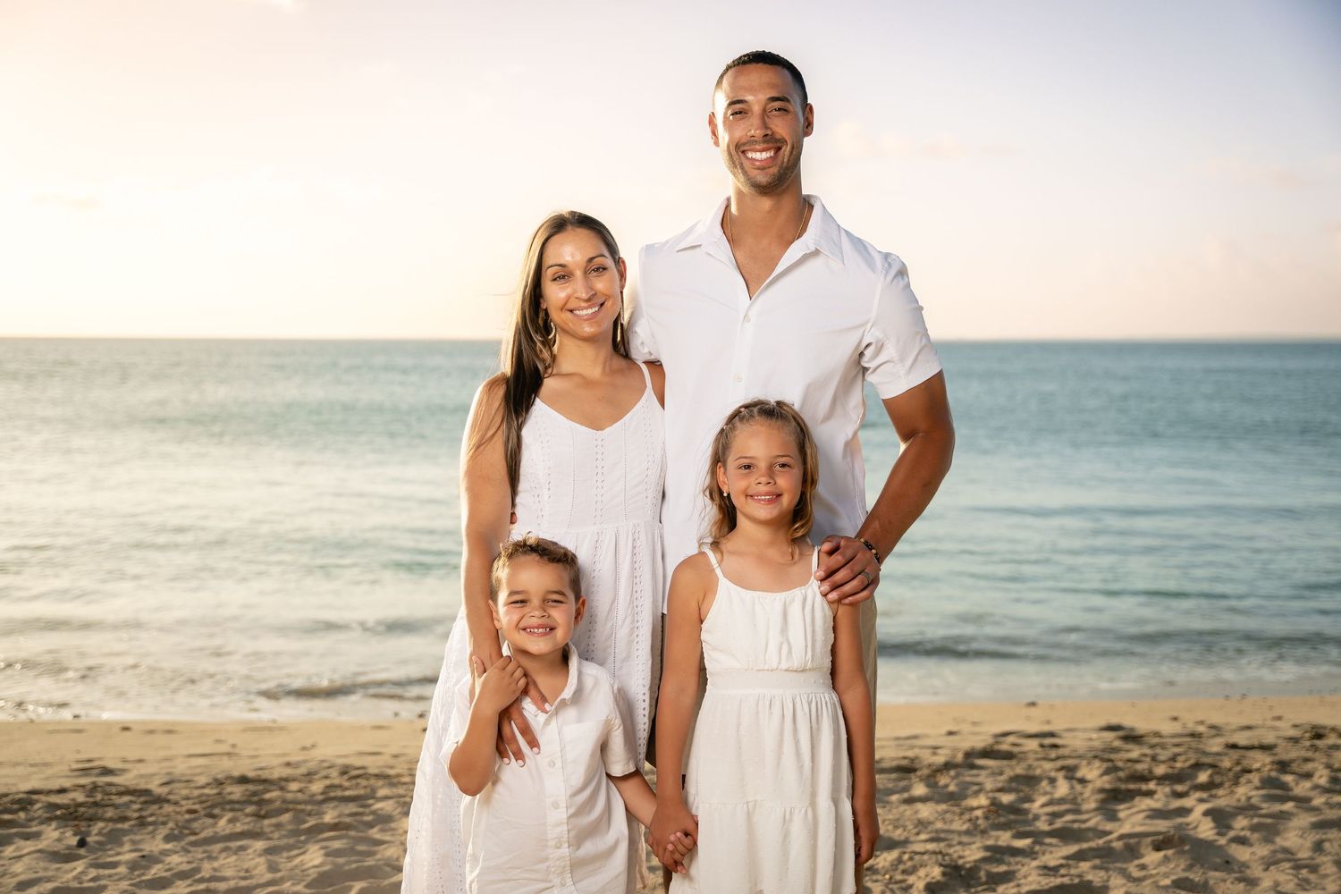 Family of four in white outfits posing on a sunny beach with ocean in background.