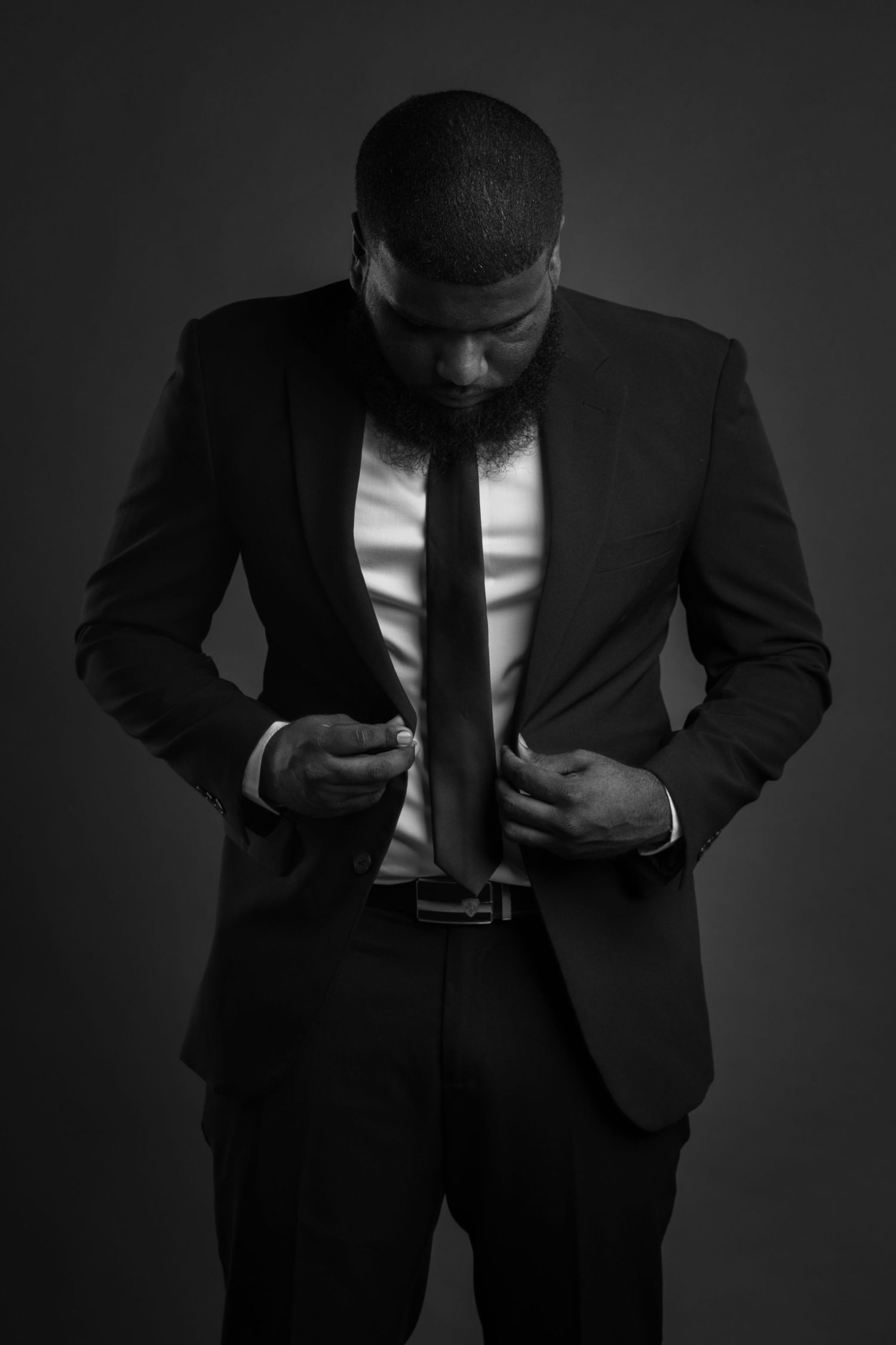 Black and white portrait of a well-dressed Black man adjusting his tie in a dark studio.