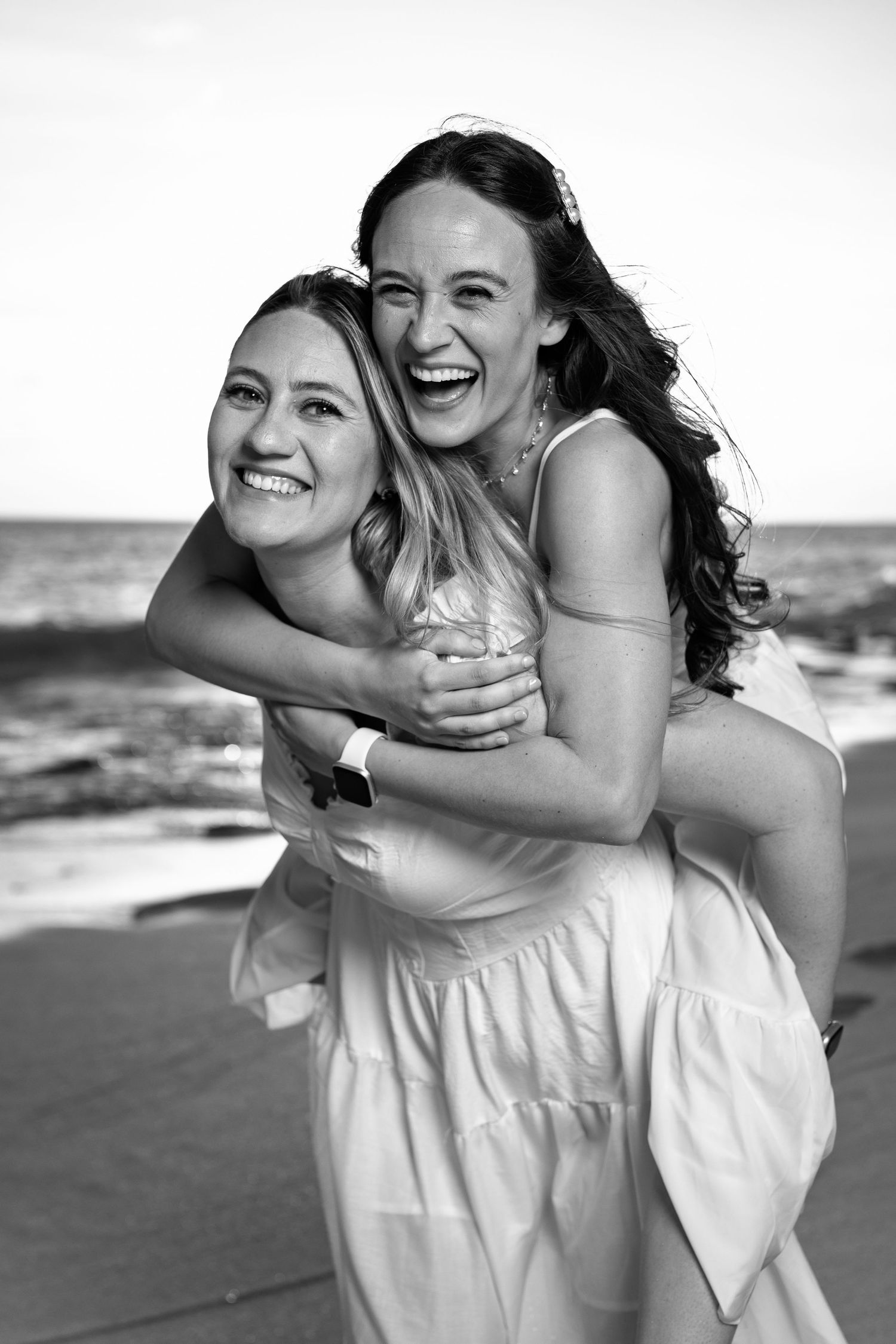 Black and white photo of two laughing women in white dresses sharing a piggyback ride at beach.