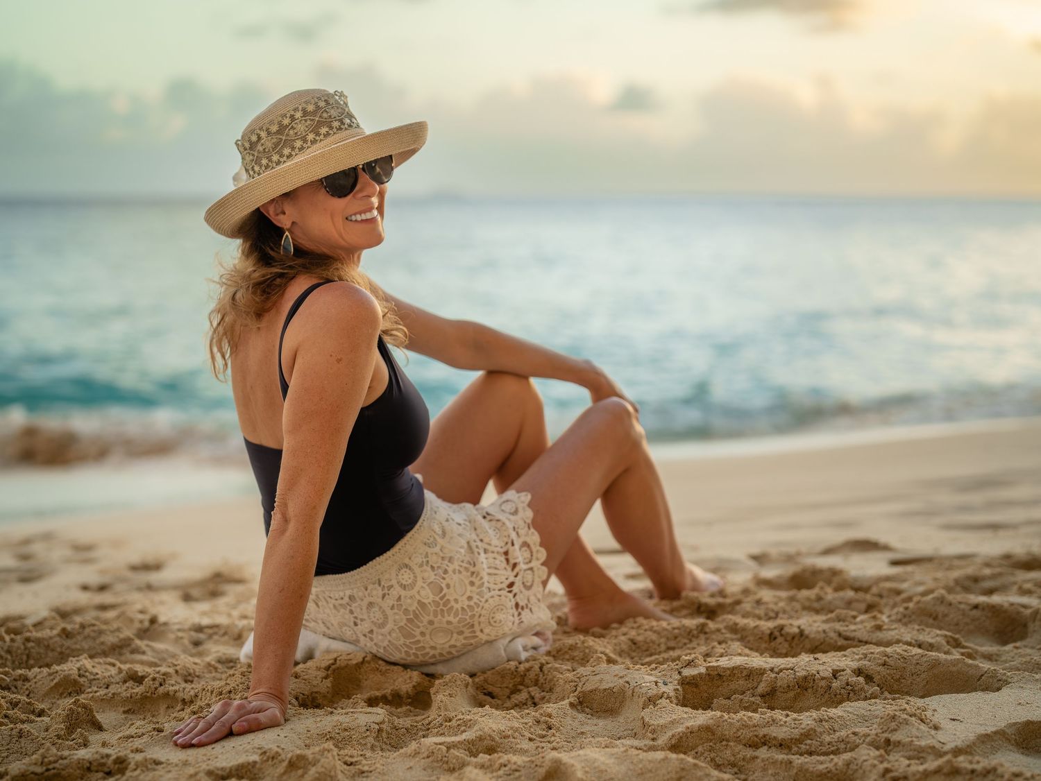 Woman in black swimsuit and straw hat sitting on sandy beach at sunset.