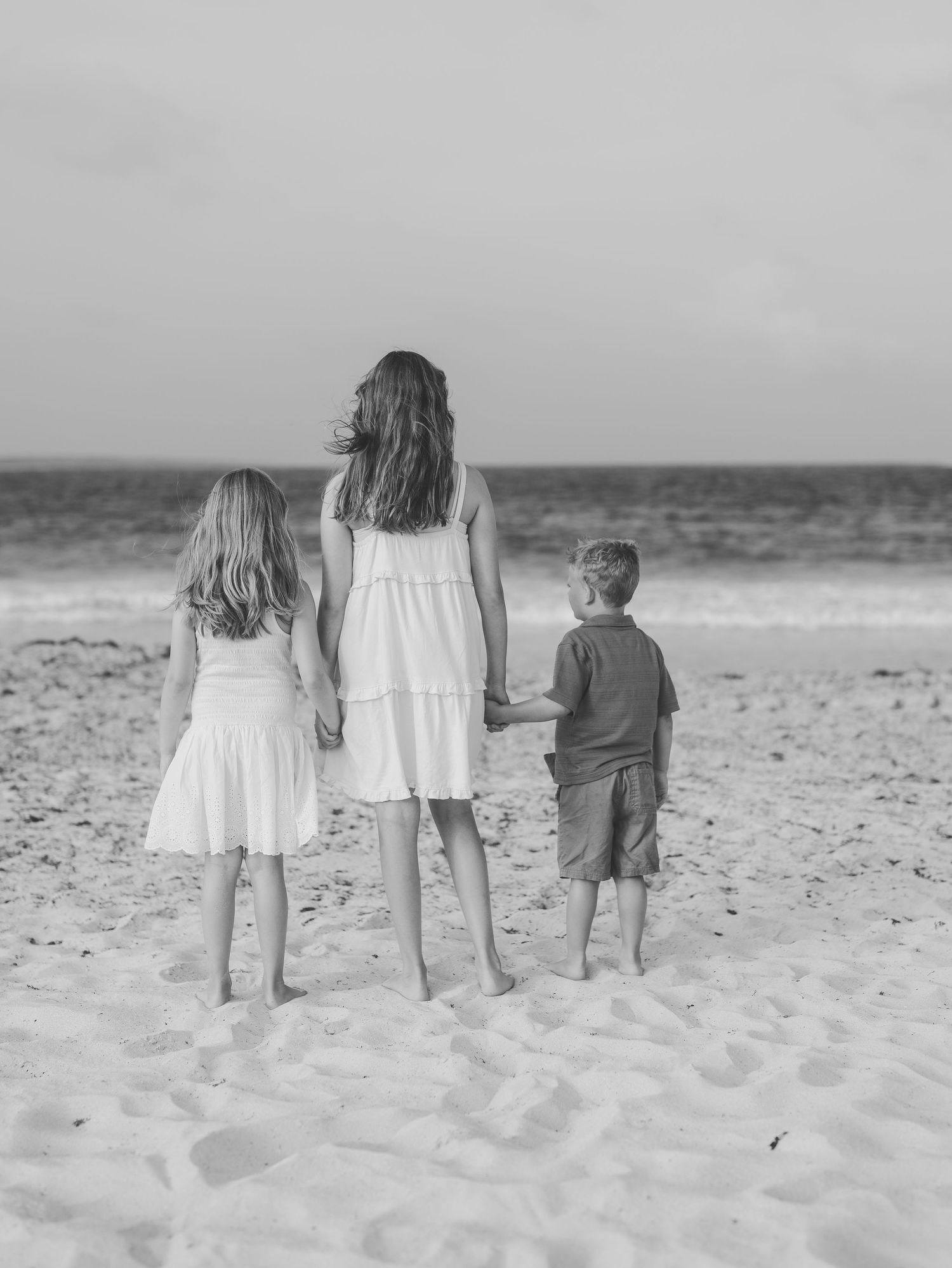 Black and white photo of three siblings holding hands facing the ocean on beach.