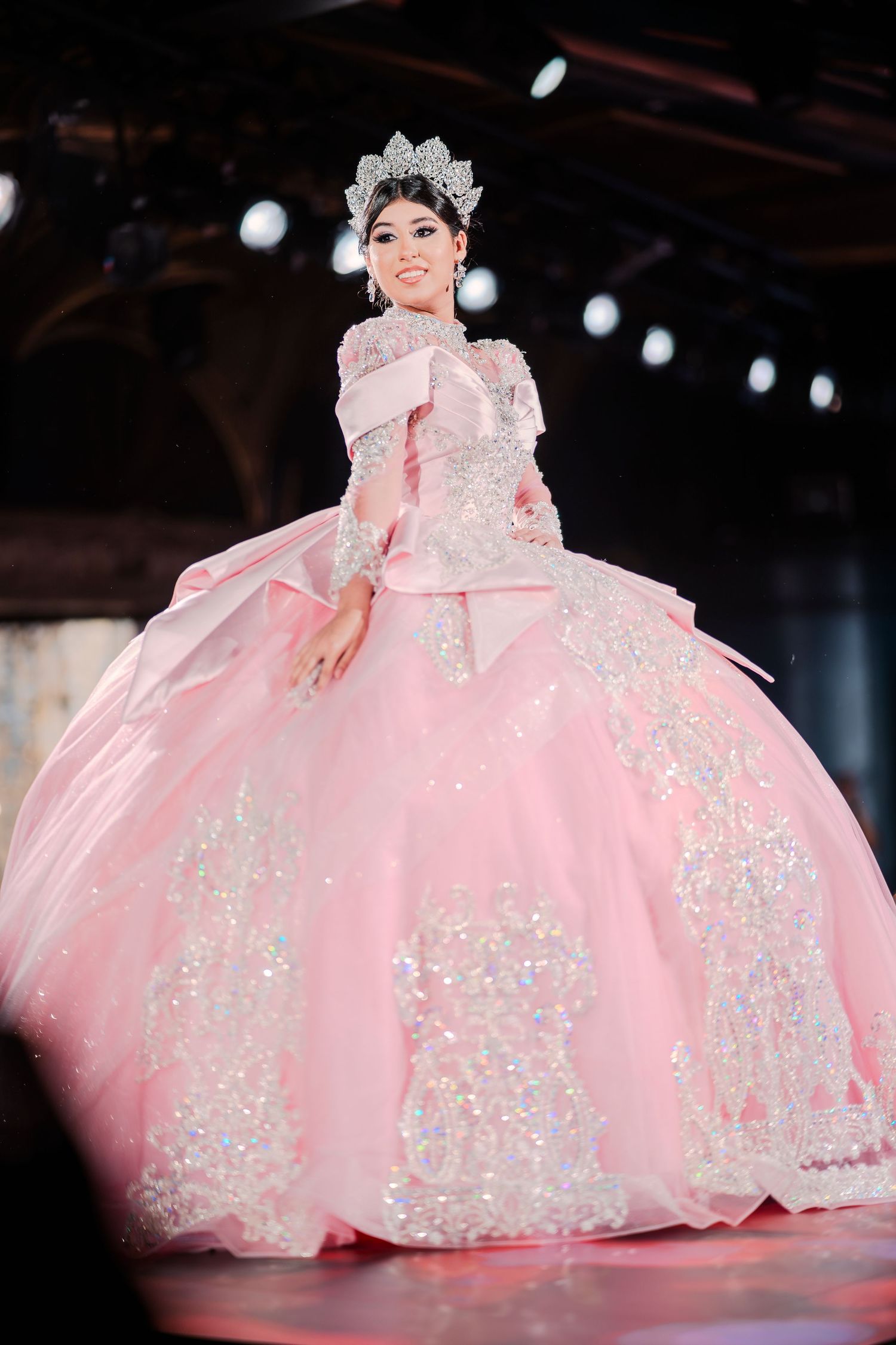 Young woman in a pink ball gown with sequins and a crystal crown poses on a runway.