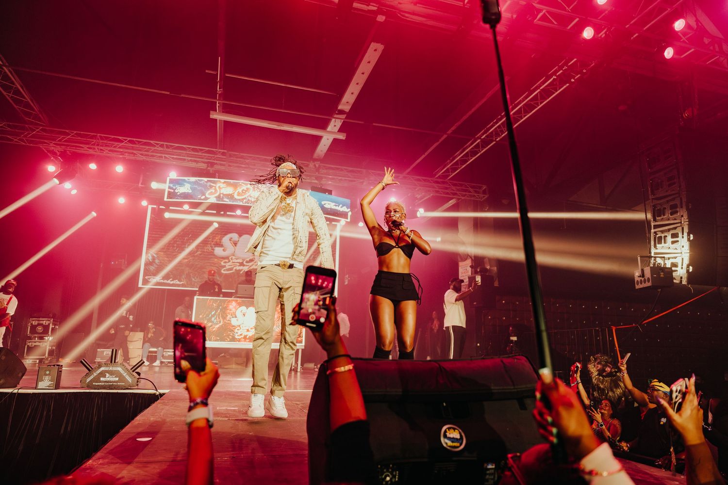 Male and female artiste performing together on a red-lit concert stage before a live crowd.