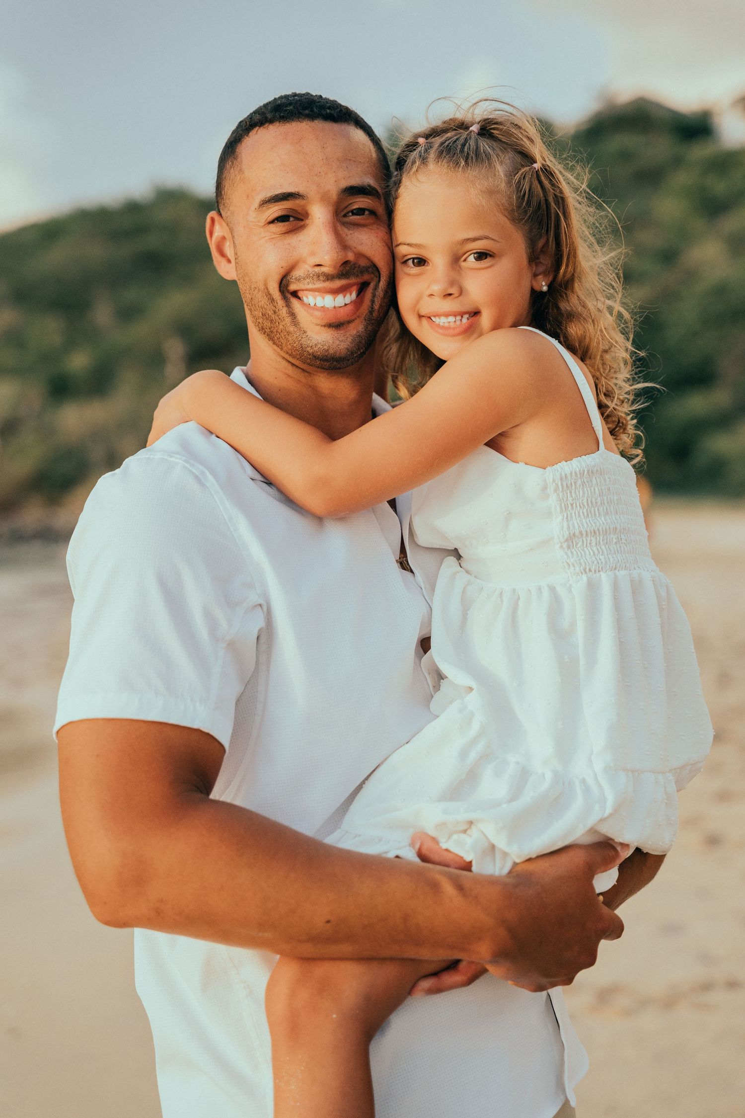Smiling father holding his curly-haired daughter in a white dress on a beach at golden hour.