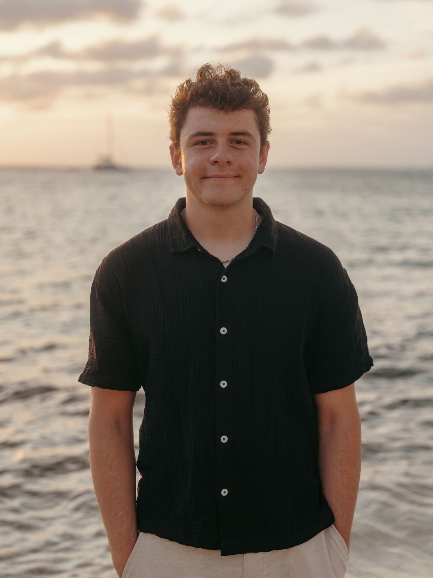 Young man in black button-up shirt smiling at camera with ocean and sailboat at sunset behind him.
