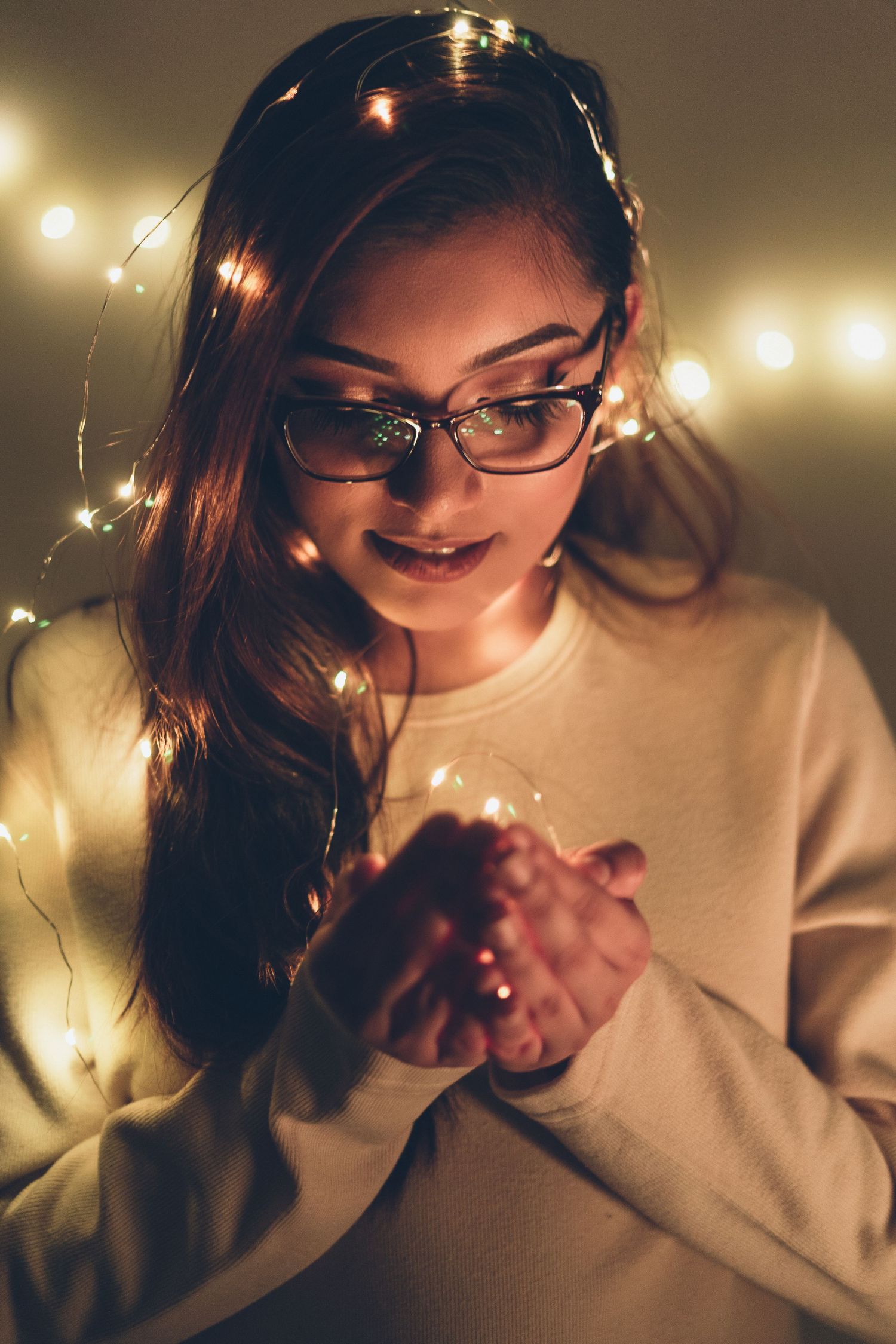 Young woman with glasses holding glowing fairy lights in a warm bokeh background.