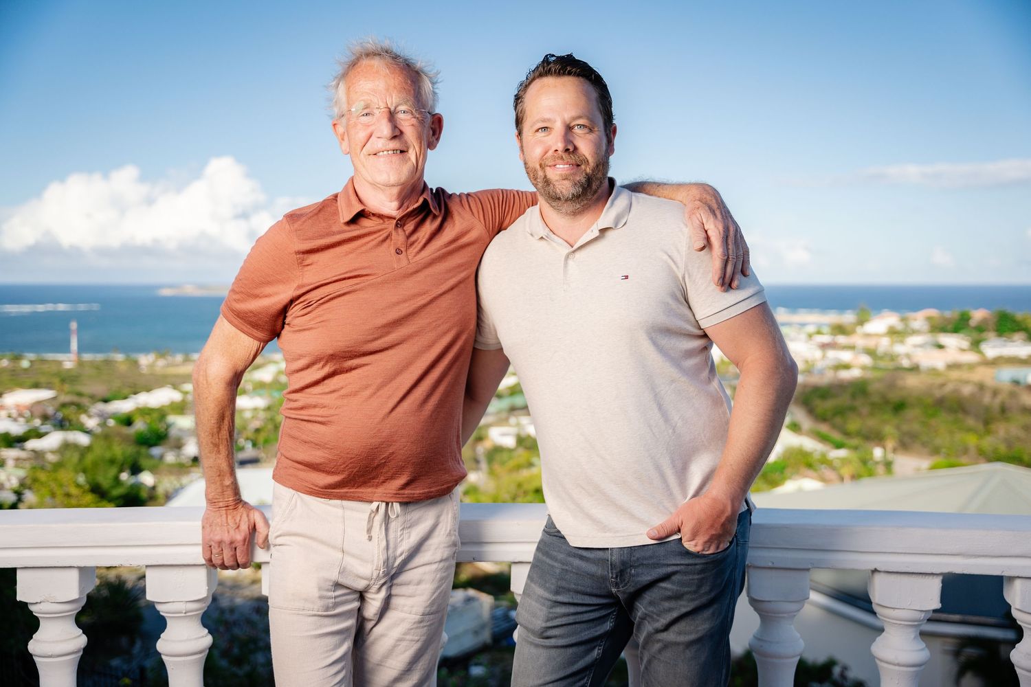 Two men smiling together on a balcony with scenic ocean view behind them.