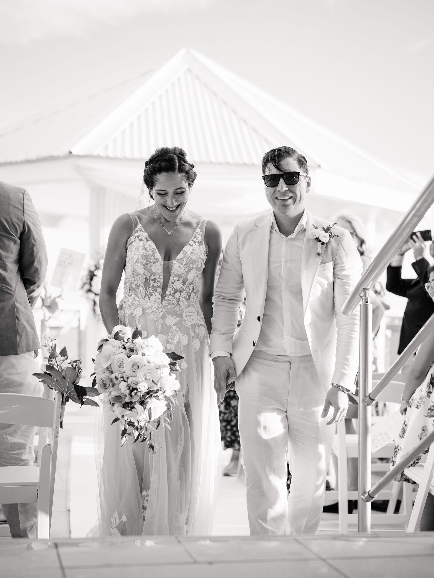 Joyful newlyweds walk down the aisle at an outdoor wedding ceremony, bride holding white bouquet.