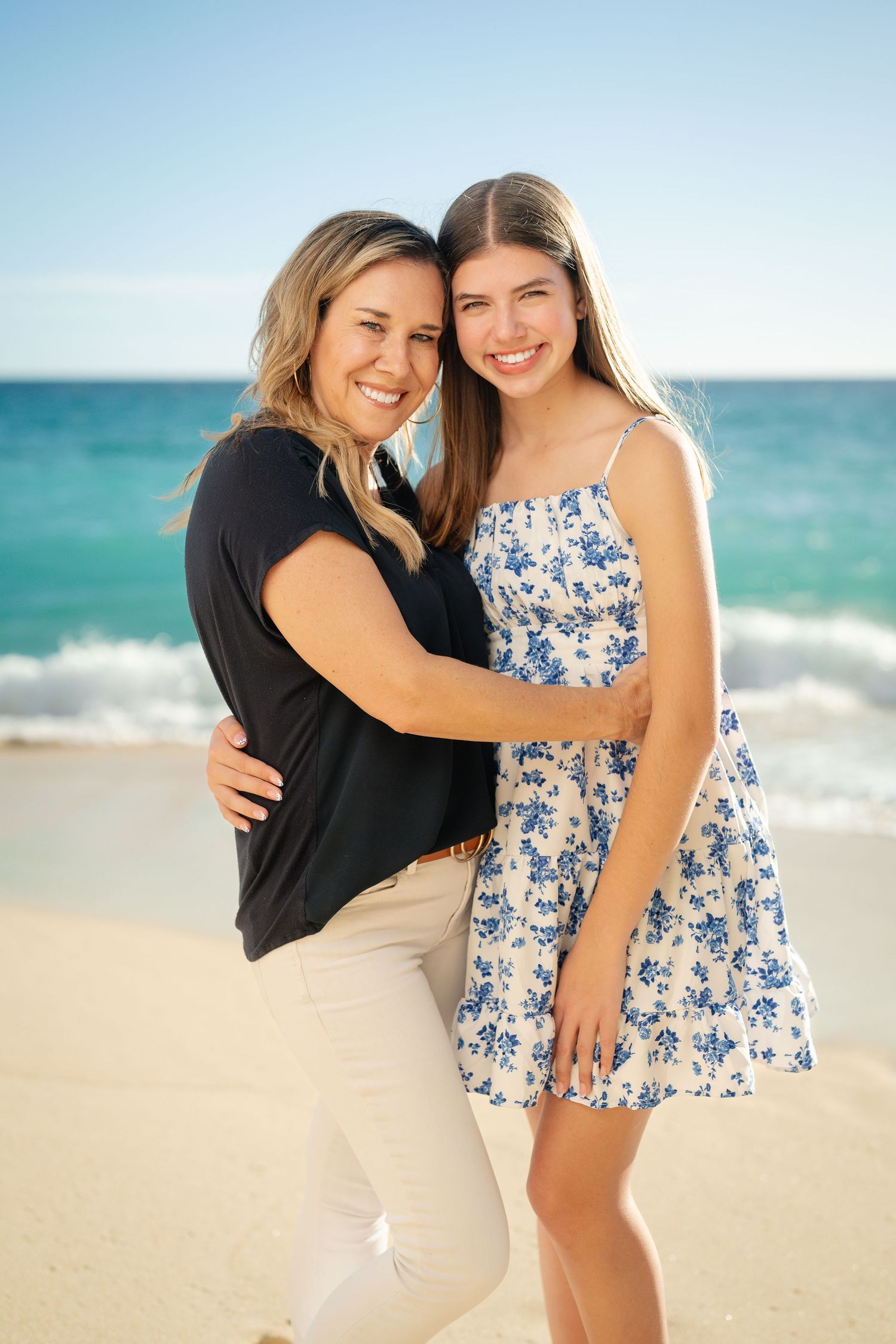 A smiling mother and teenage daughter embrace on a sunny beach with turquoise ocean waves behind them.
