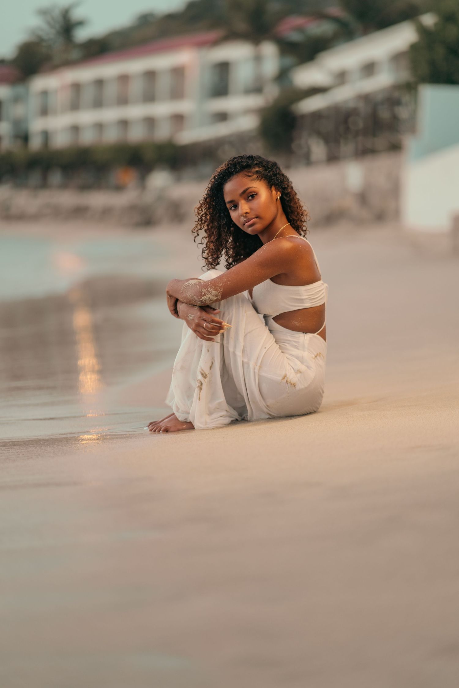 Young woman with curly hair in white outfit sitting on sandy beach at sunset near resort buildings.