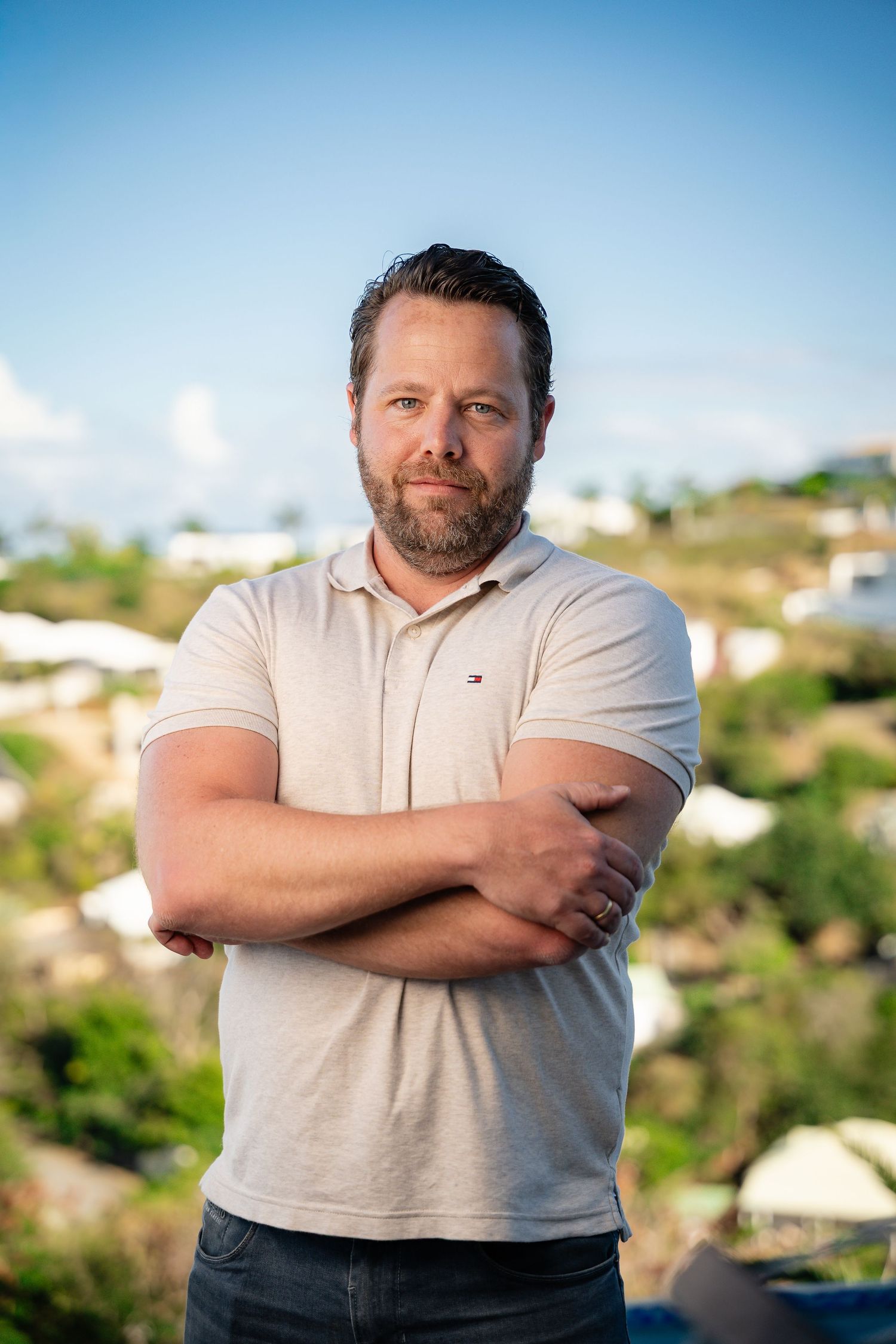 Man with beard wearing a Tommy Hilfiger polo shirt standing with arms crossed outdoors.