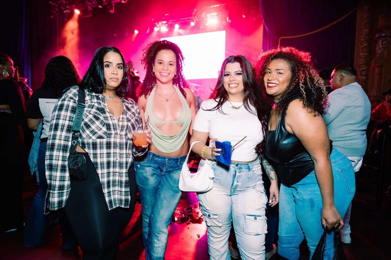 Four women smiling and posing together at a lively indoor concert event.