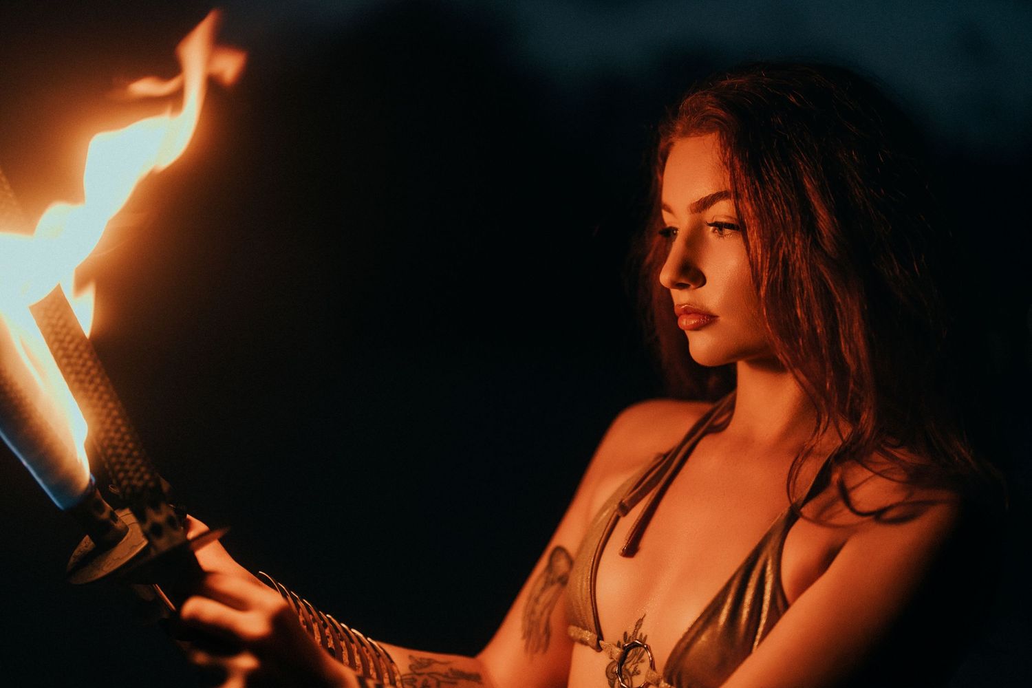 A tattooed young woman holds a flaming torch in the dark during a nighttime fire performance.