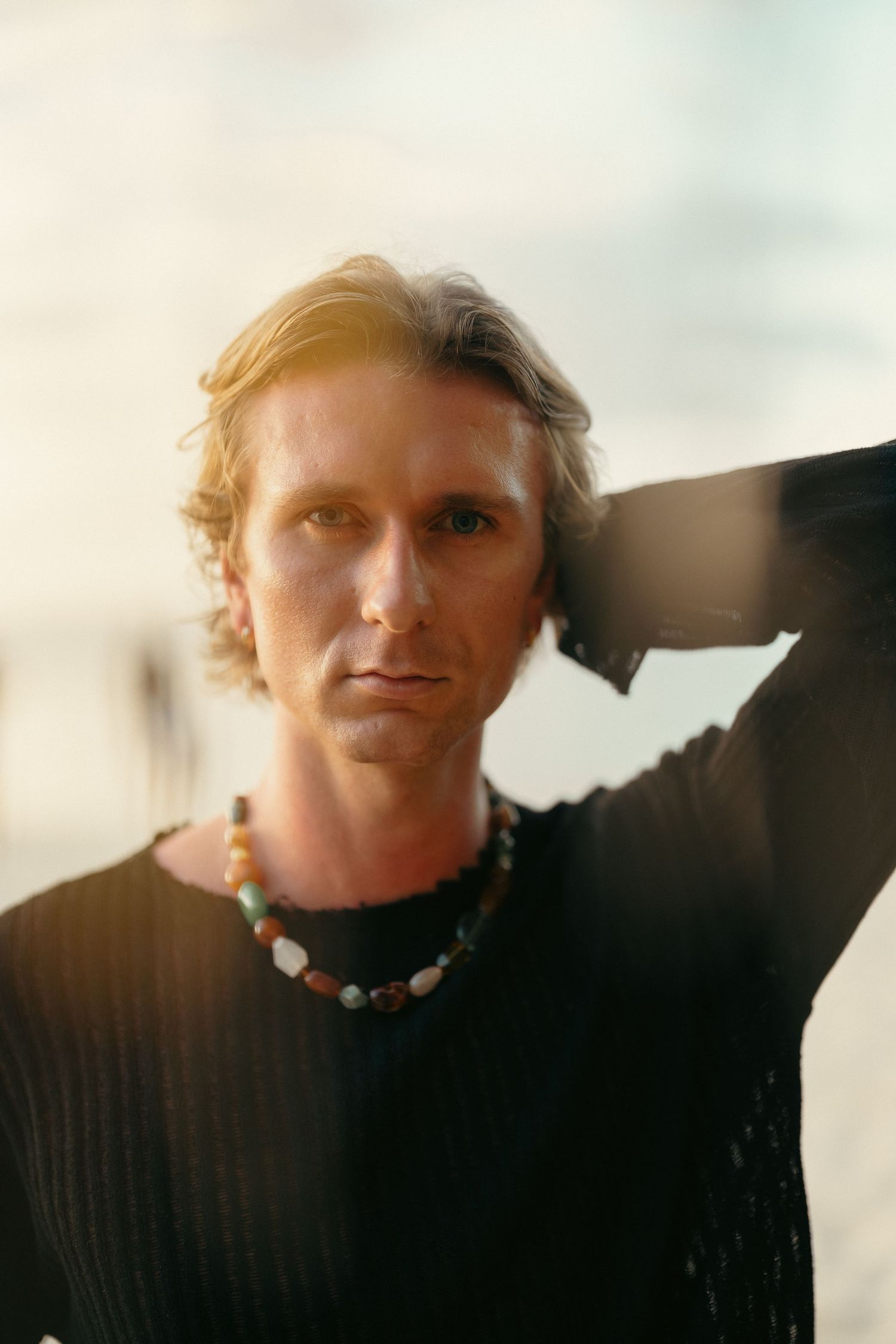 Blond man in black sweater wearing beaded necklace in soft sunlit outdoor portrait.