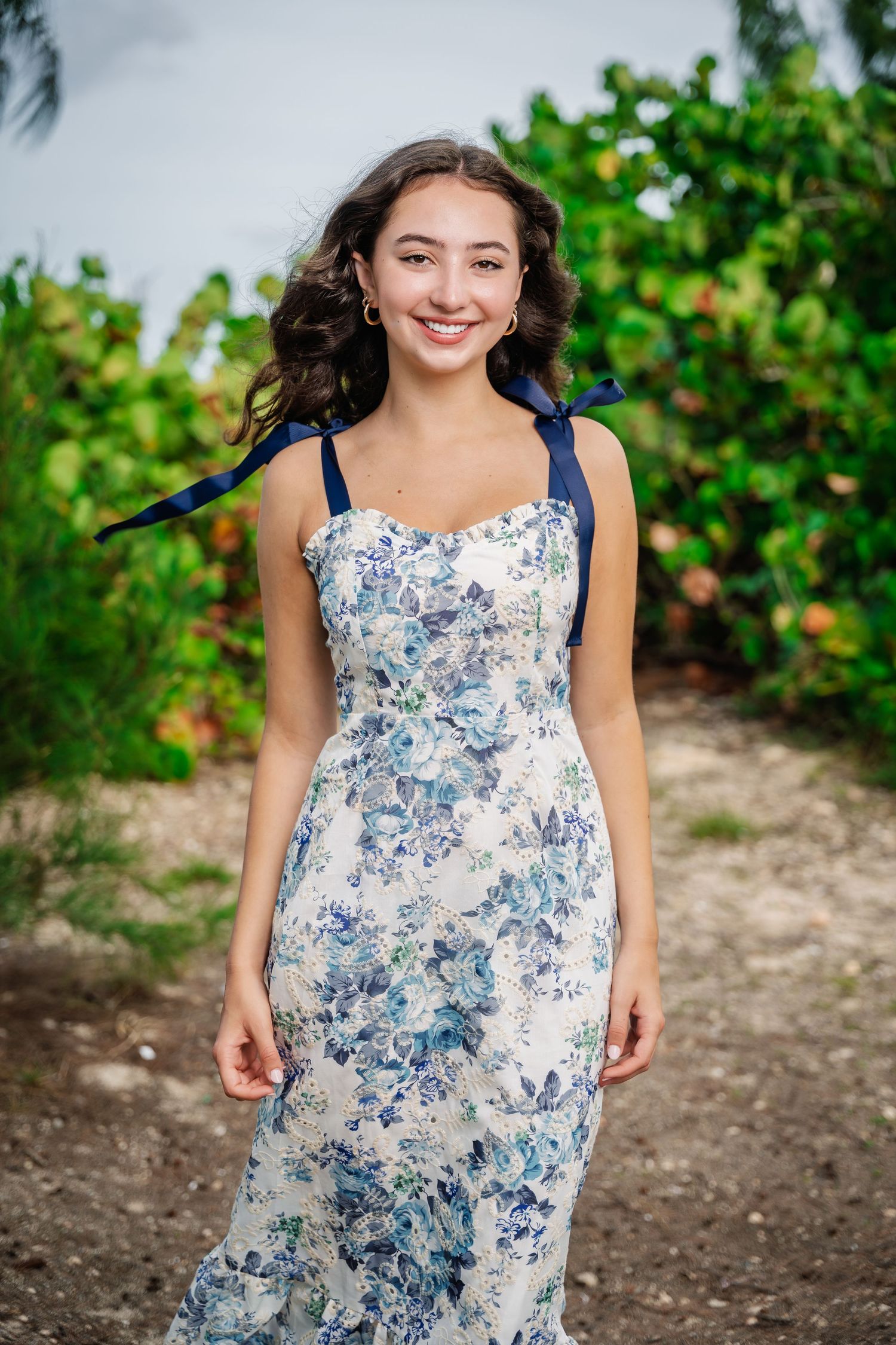 Smiling young woman in a blue floral midi dress standing on a dirt path surrounded by lush greenery.