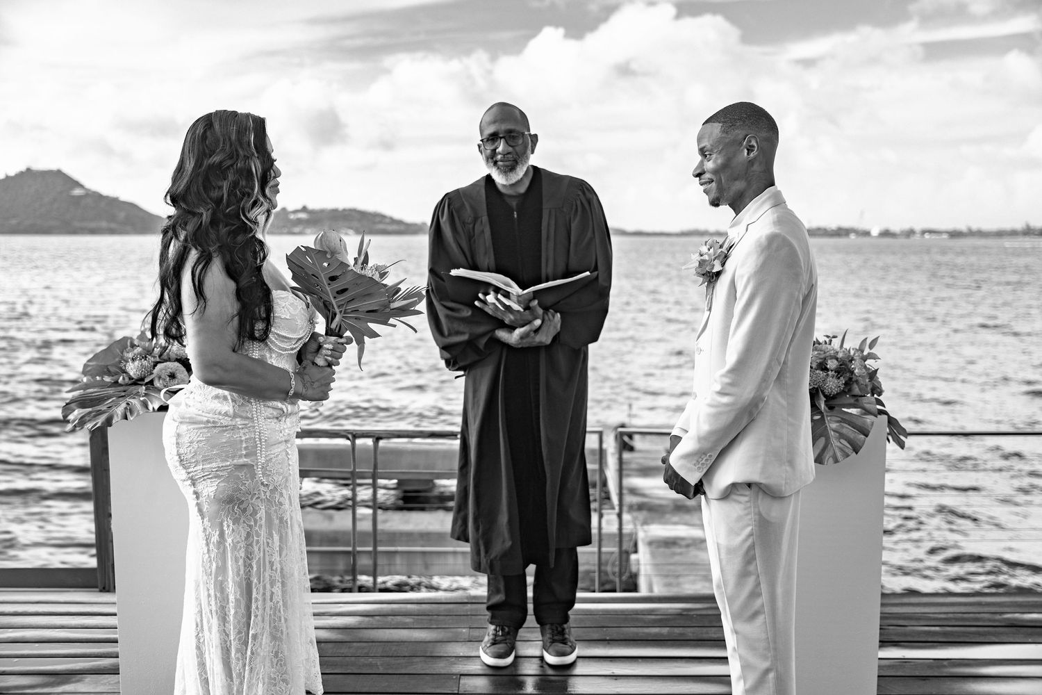 Black and white photo of couple exchanging vows on dock with officiant and ocean backdrop.