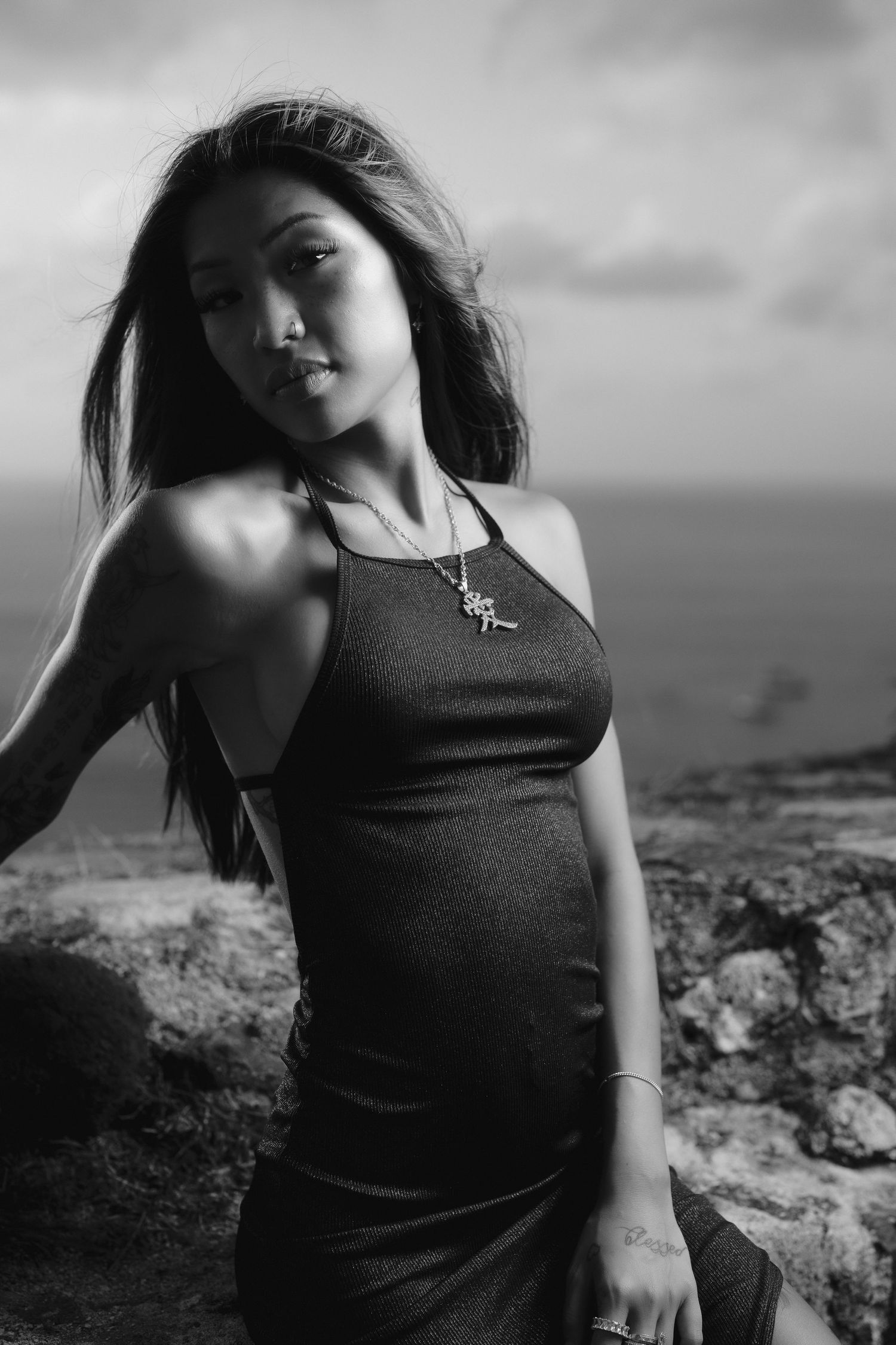 Black and white portrait of young Asian woman in sleeveless dress with necklace by ocean.