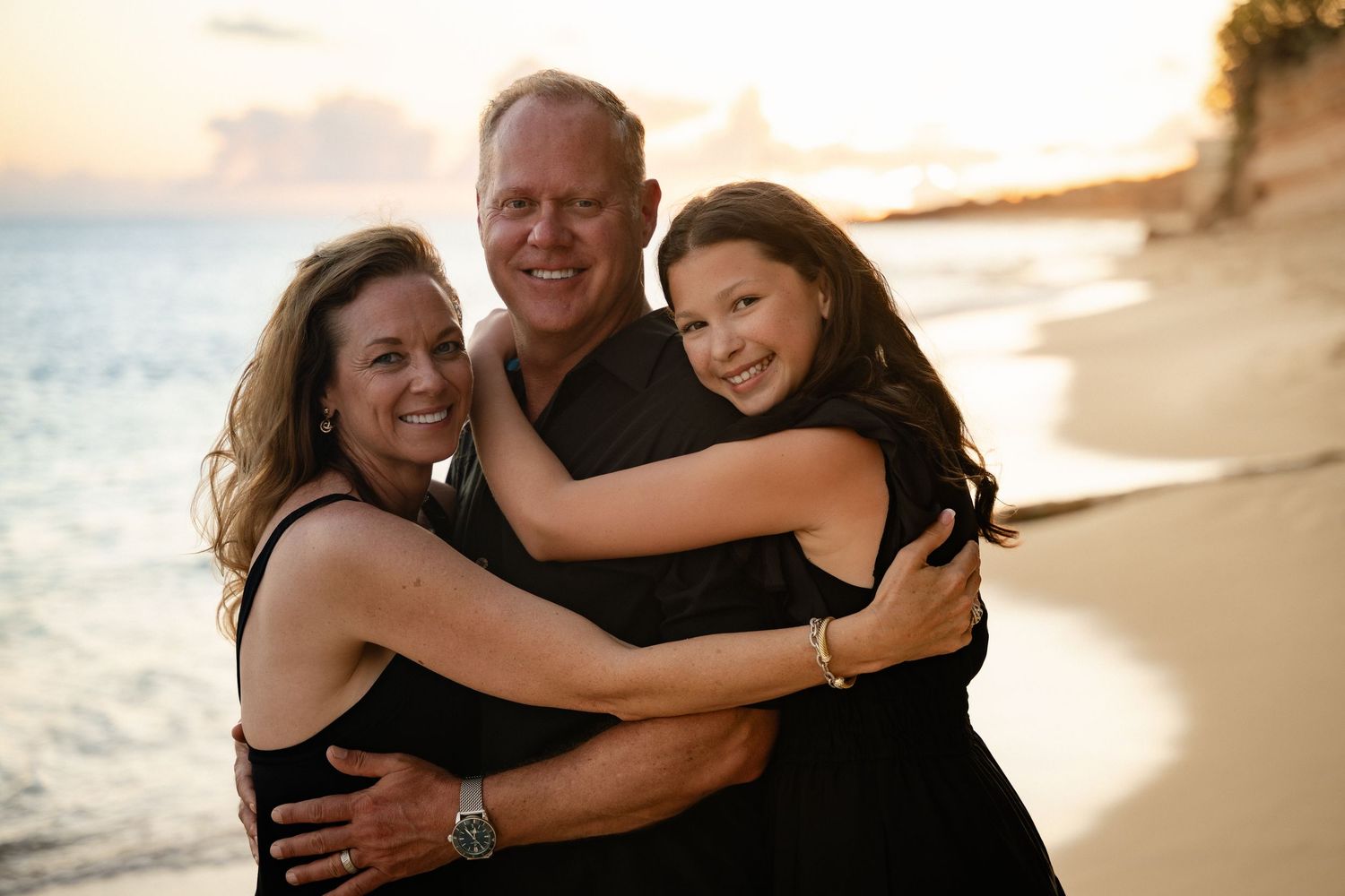 Three family members hugging and smiling on a beach at sunset.