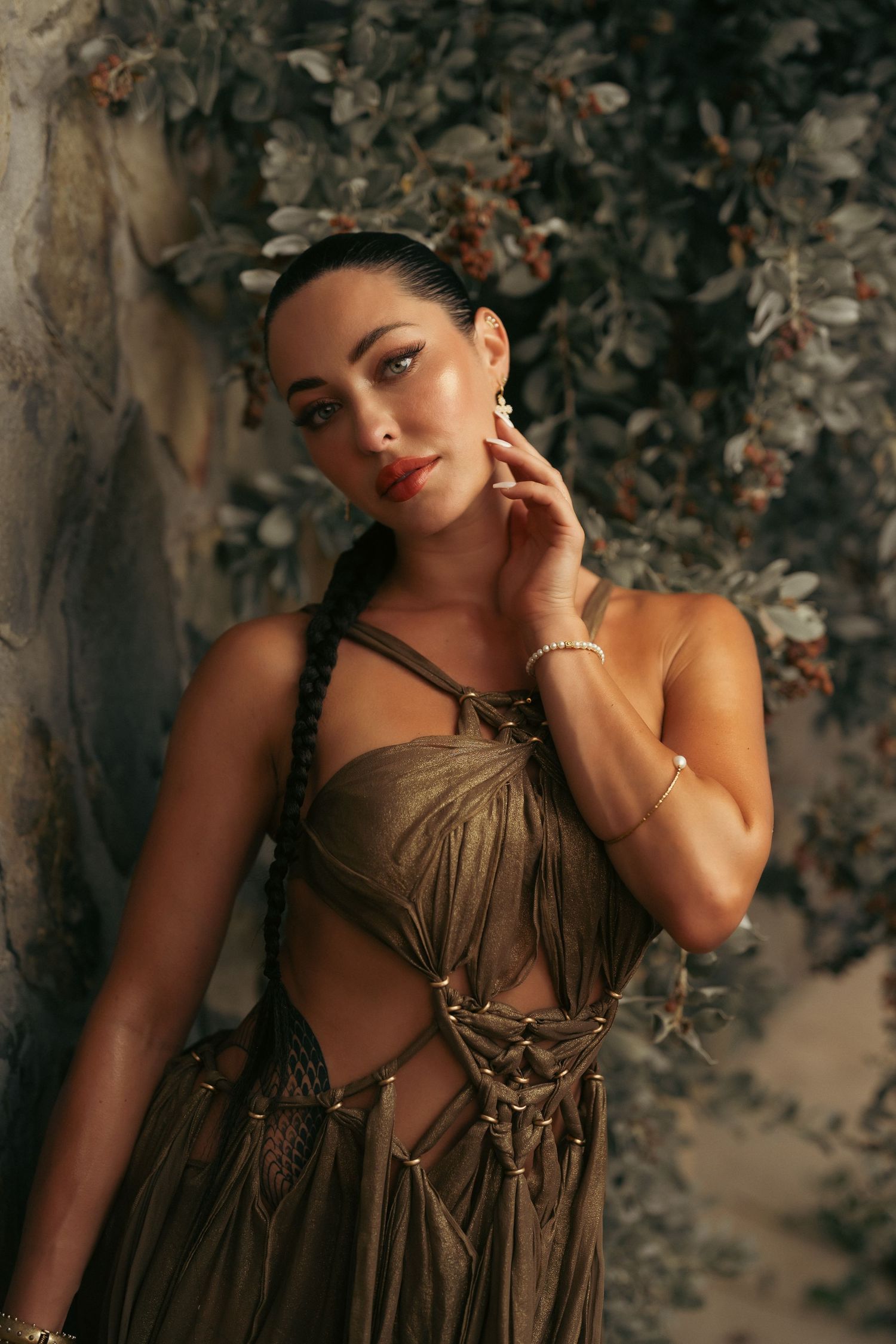 Woman with a side braid wearing a gold macrame dress posing against a stone wall with foliage backdrop.