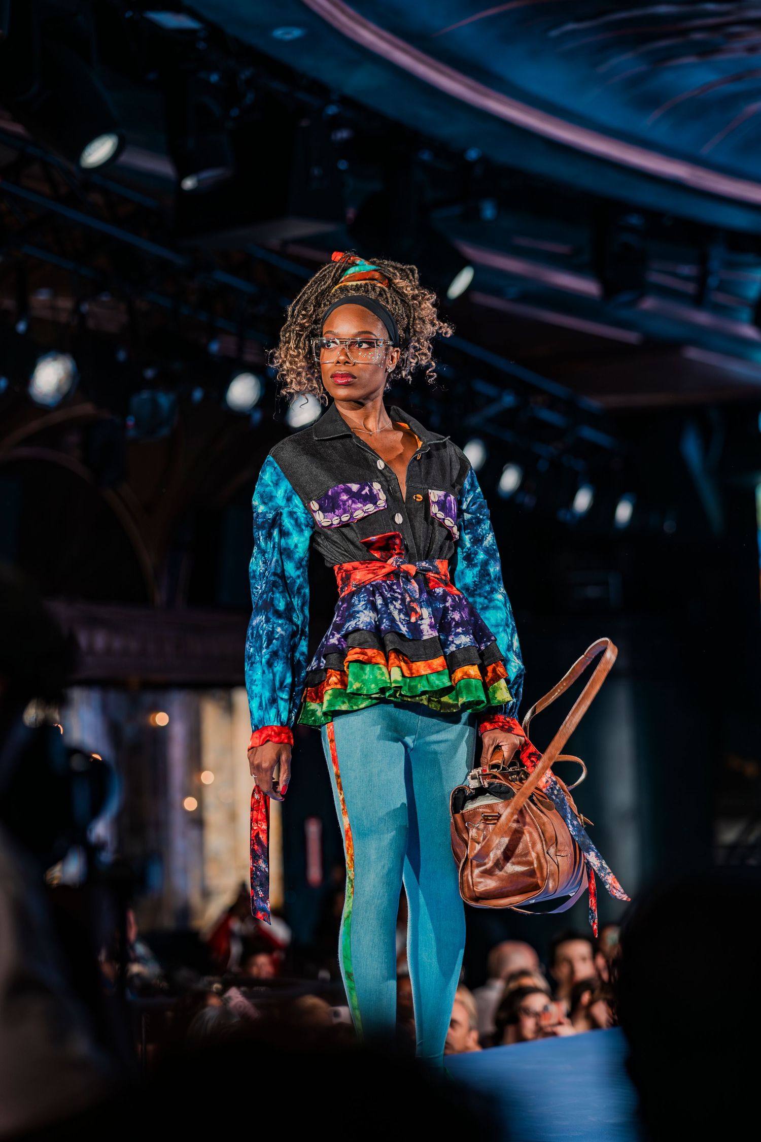 A model walks the runway in a vibrant multicolor patchwork jacket, teal tights, and carries a brown leather bag.