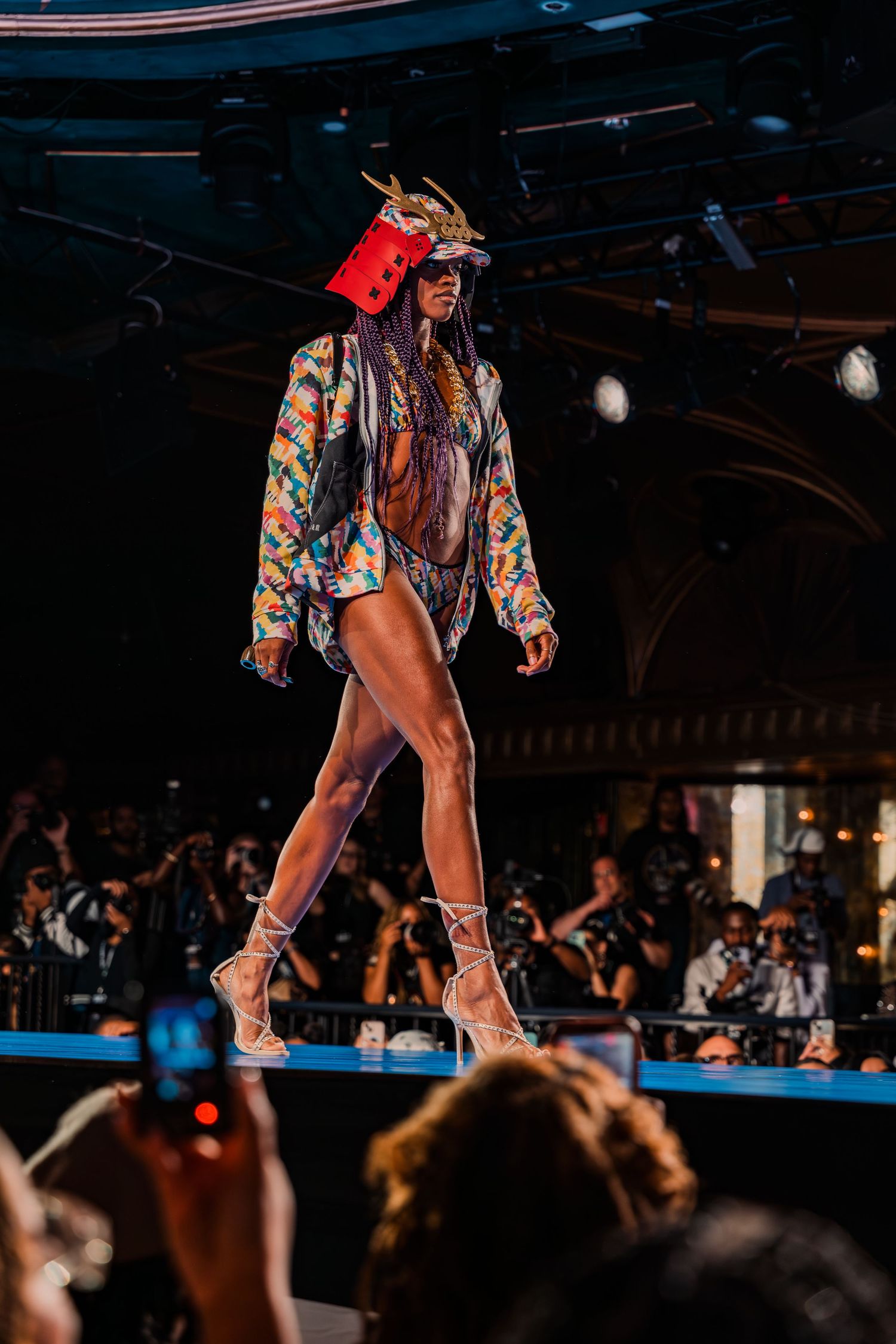 Model walks runway in colorful jacket, bold headpiece, and strappy heels at a vibrant fashion show.