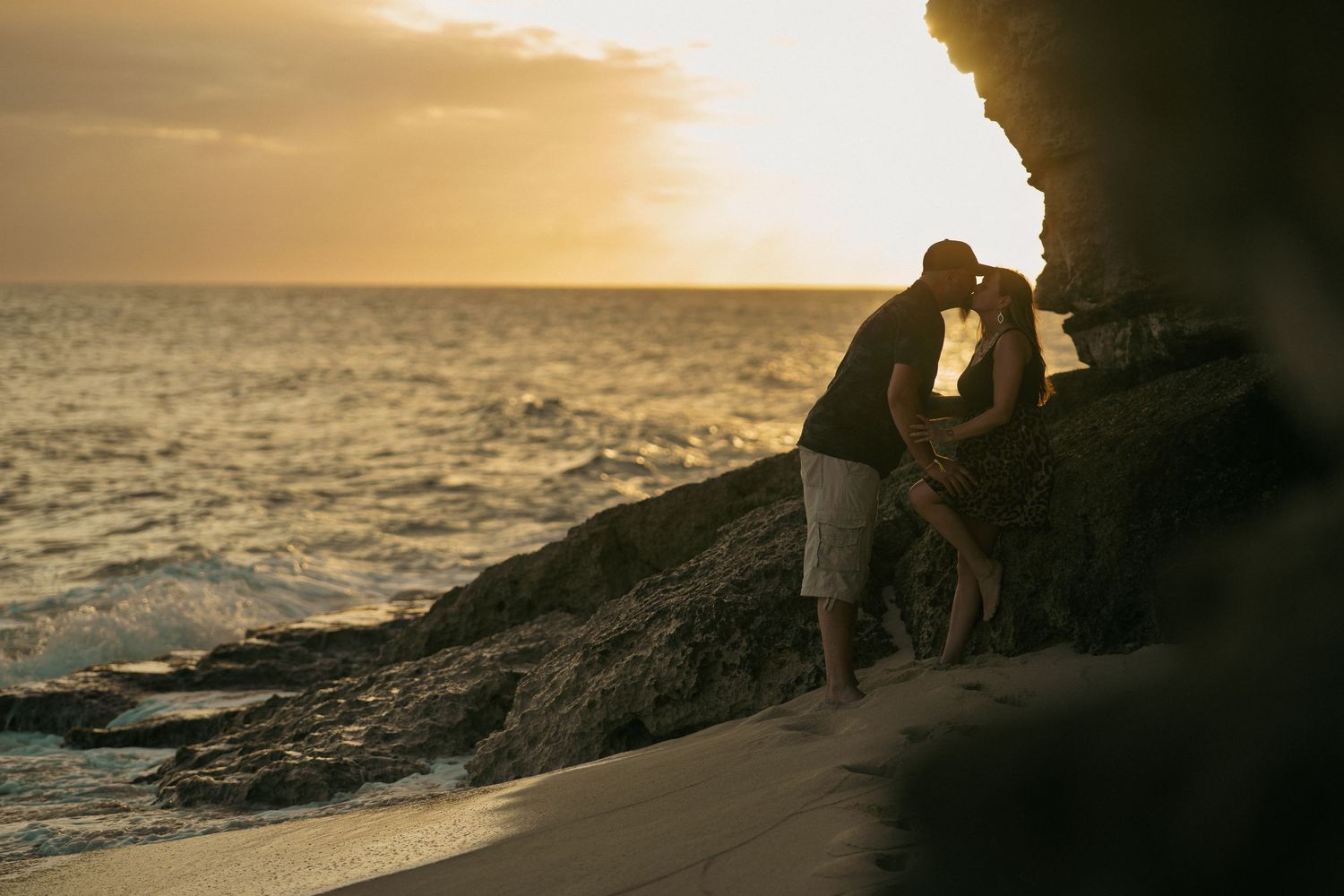 Couple sharing romantic kiss against rocky cliff at golden sunset on beach.