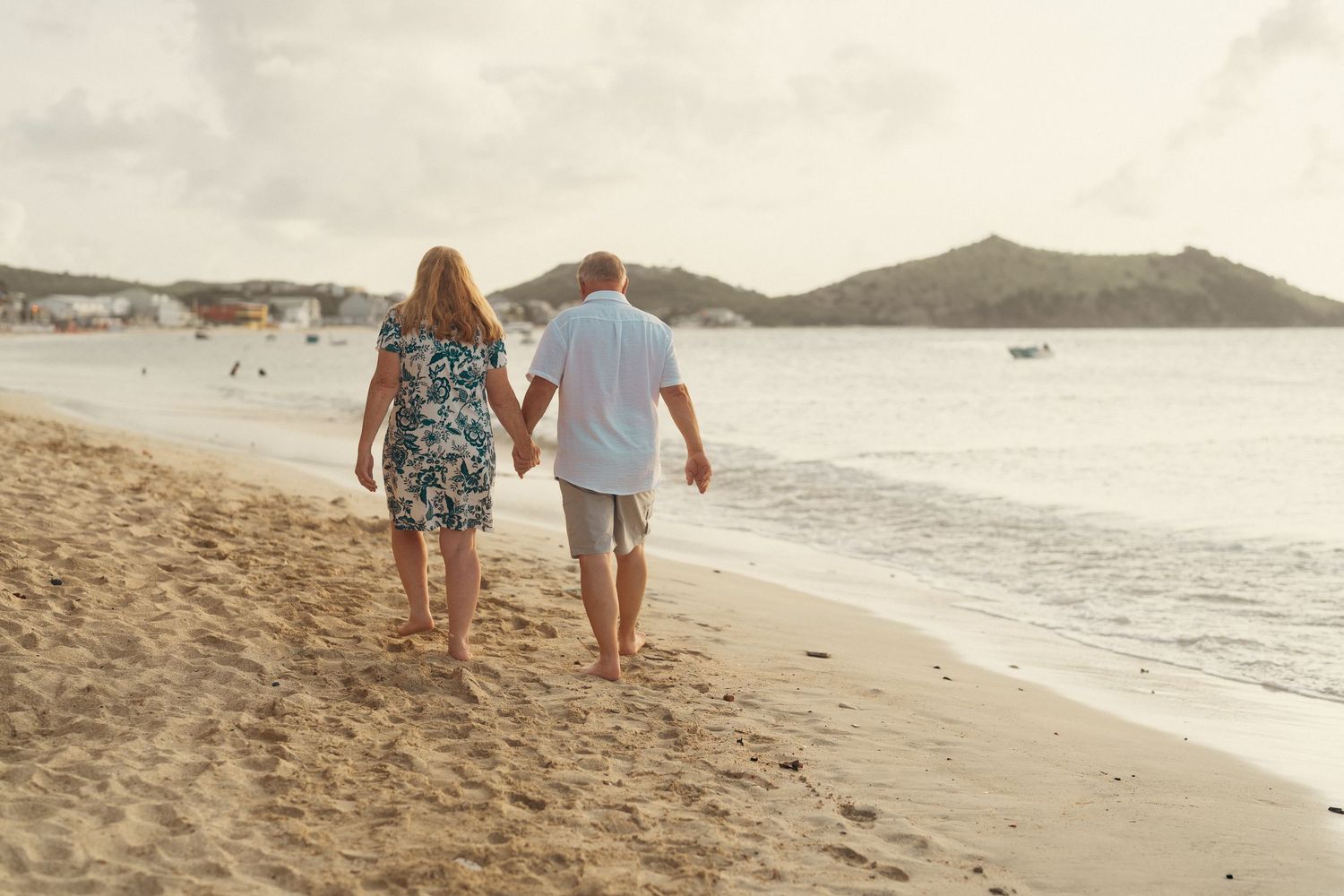 A couple walks hand-in-hand along a tropical beach at sunset with mountains visible in the distance.