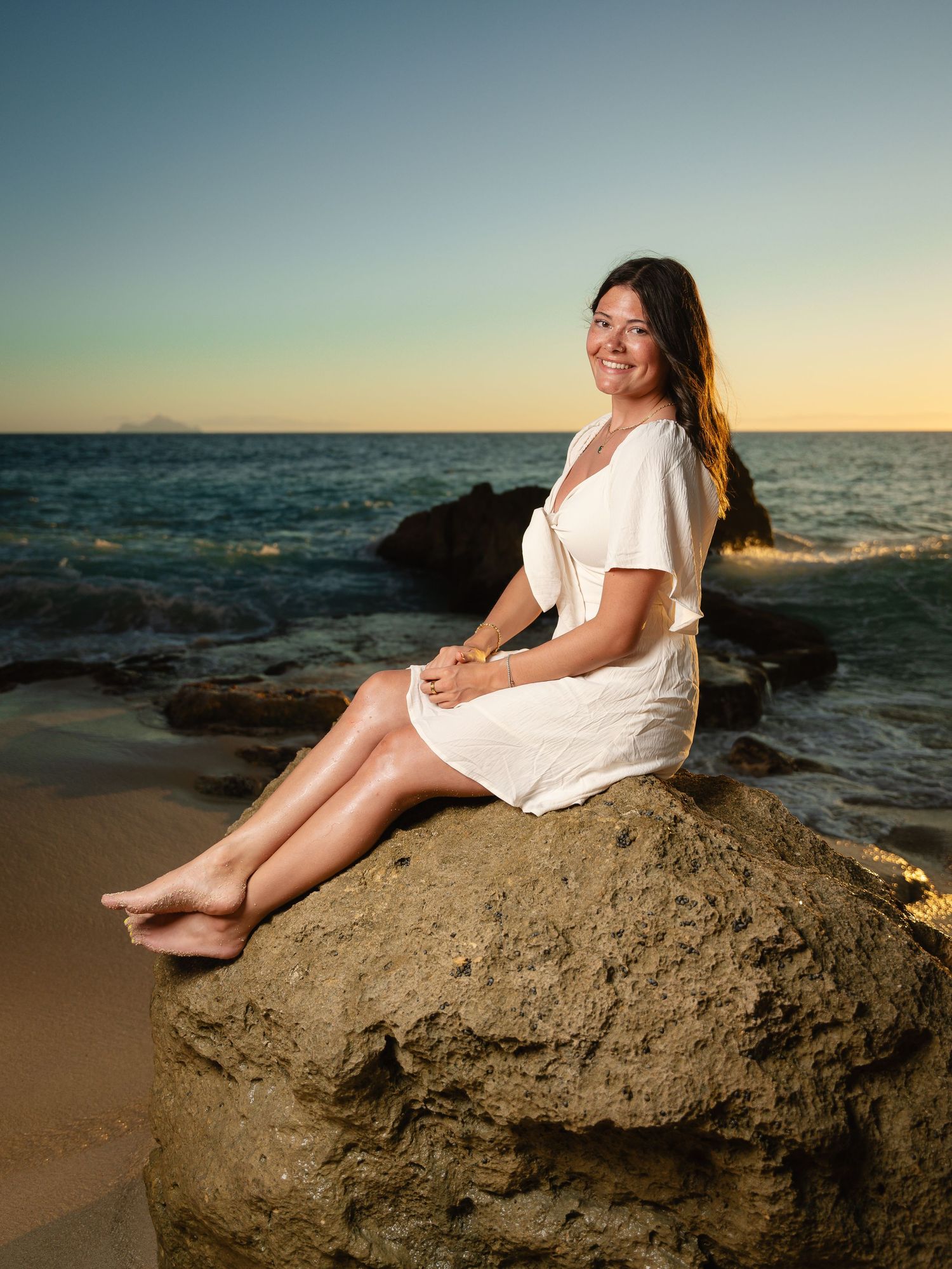 Smiling young woman in white dress sitting barefoot on a rock at a scenic beach during sunset.