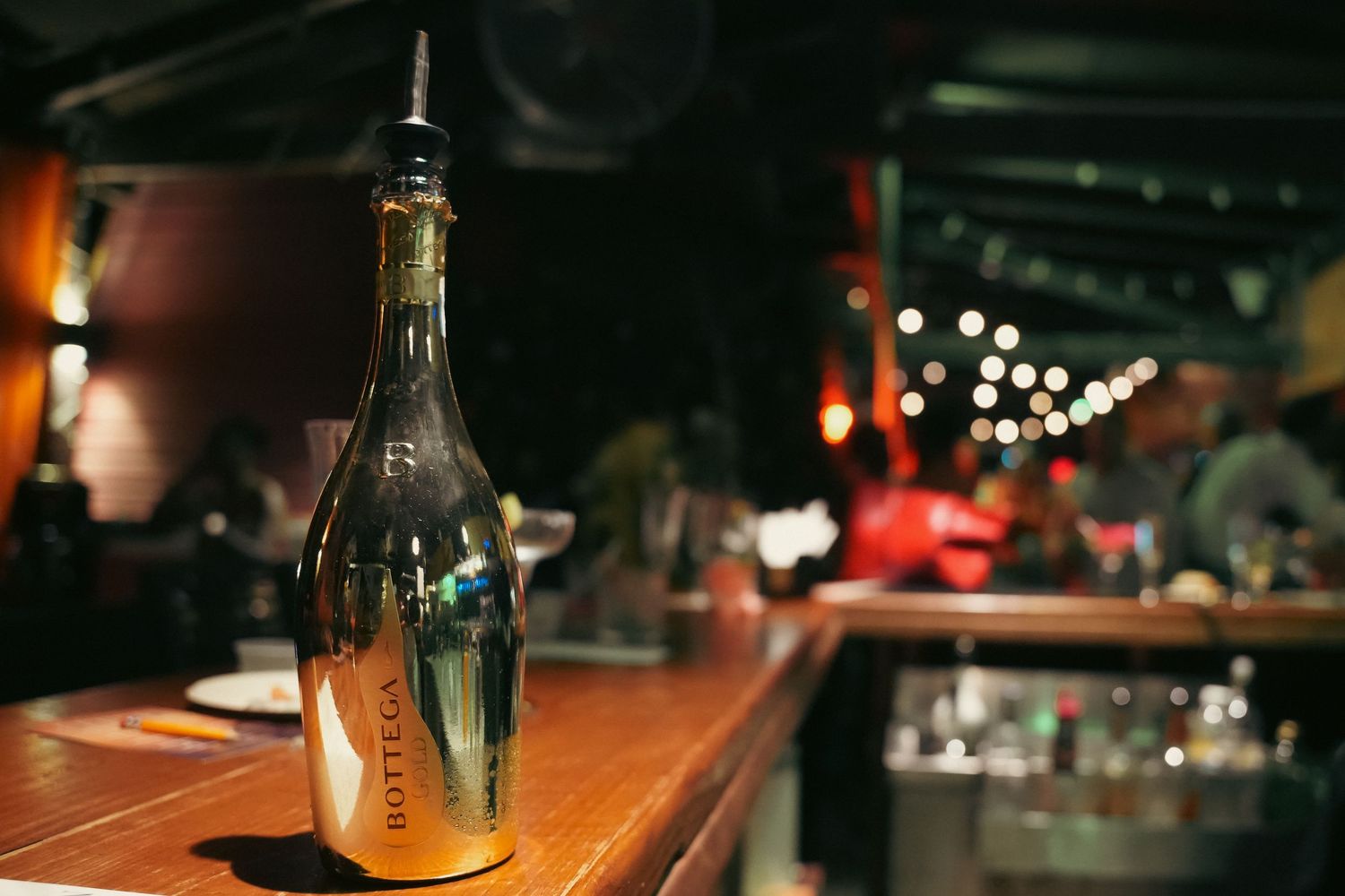 Gold wine bottle on a wooden bar counter with bokeh lights in a dark restaurant.
