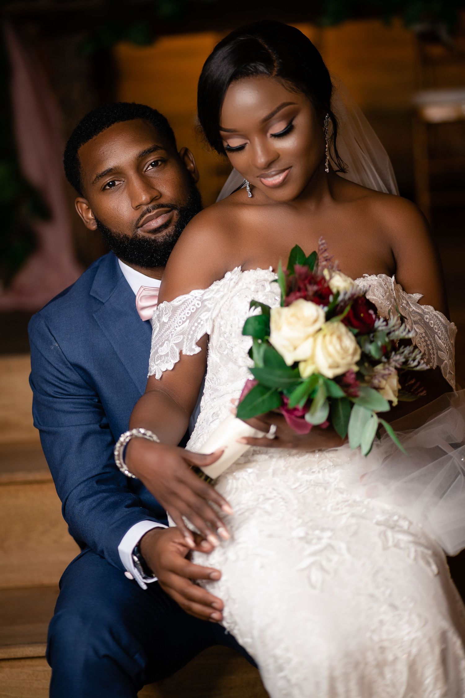 Groom in navy suit embraces bride in lace gown holding colorful bouquet at wedding.
